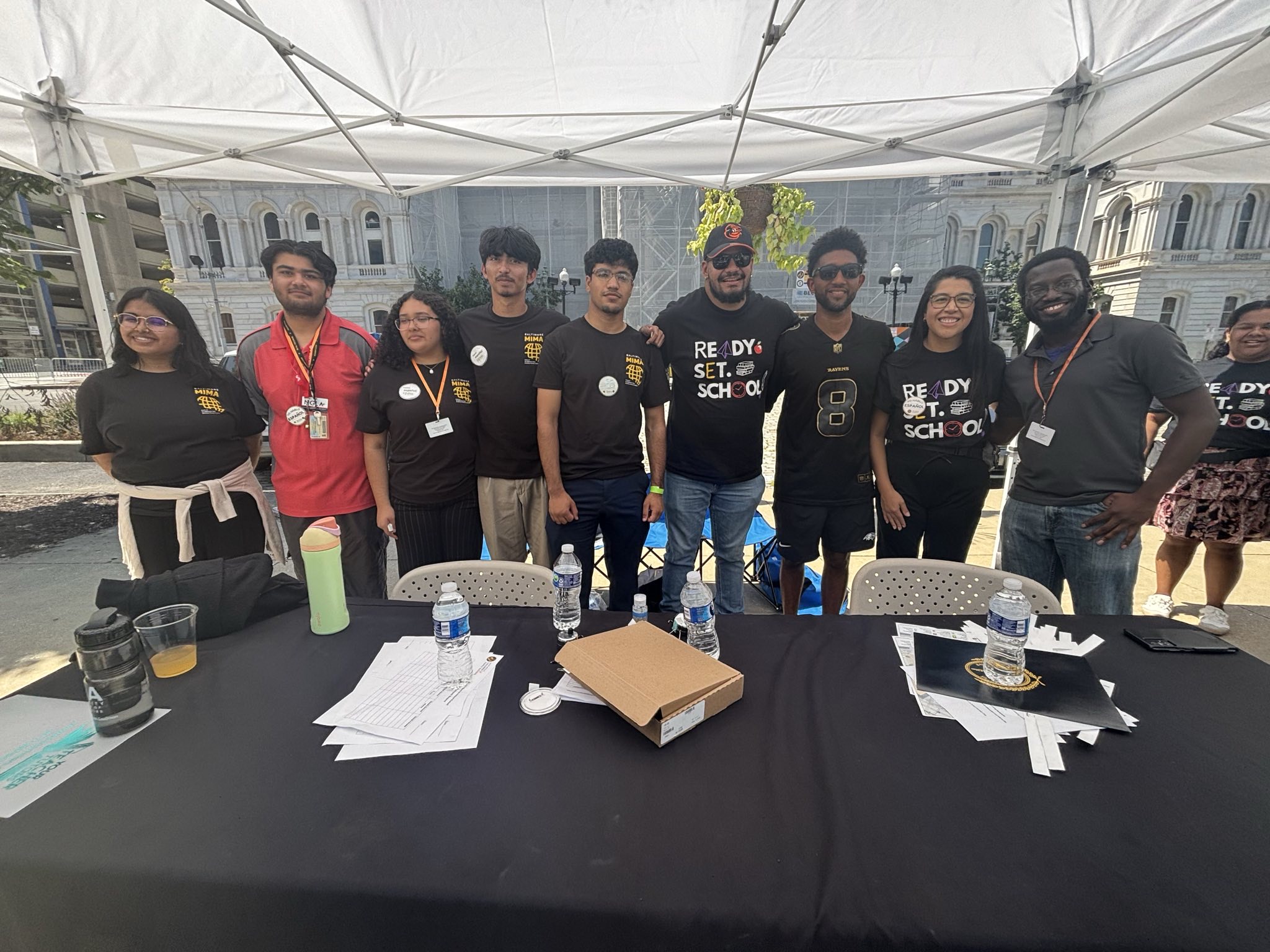 Group of nine people standing behind a table with papers and water bottles, under a white canopy outdoors.