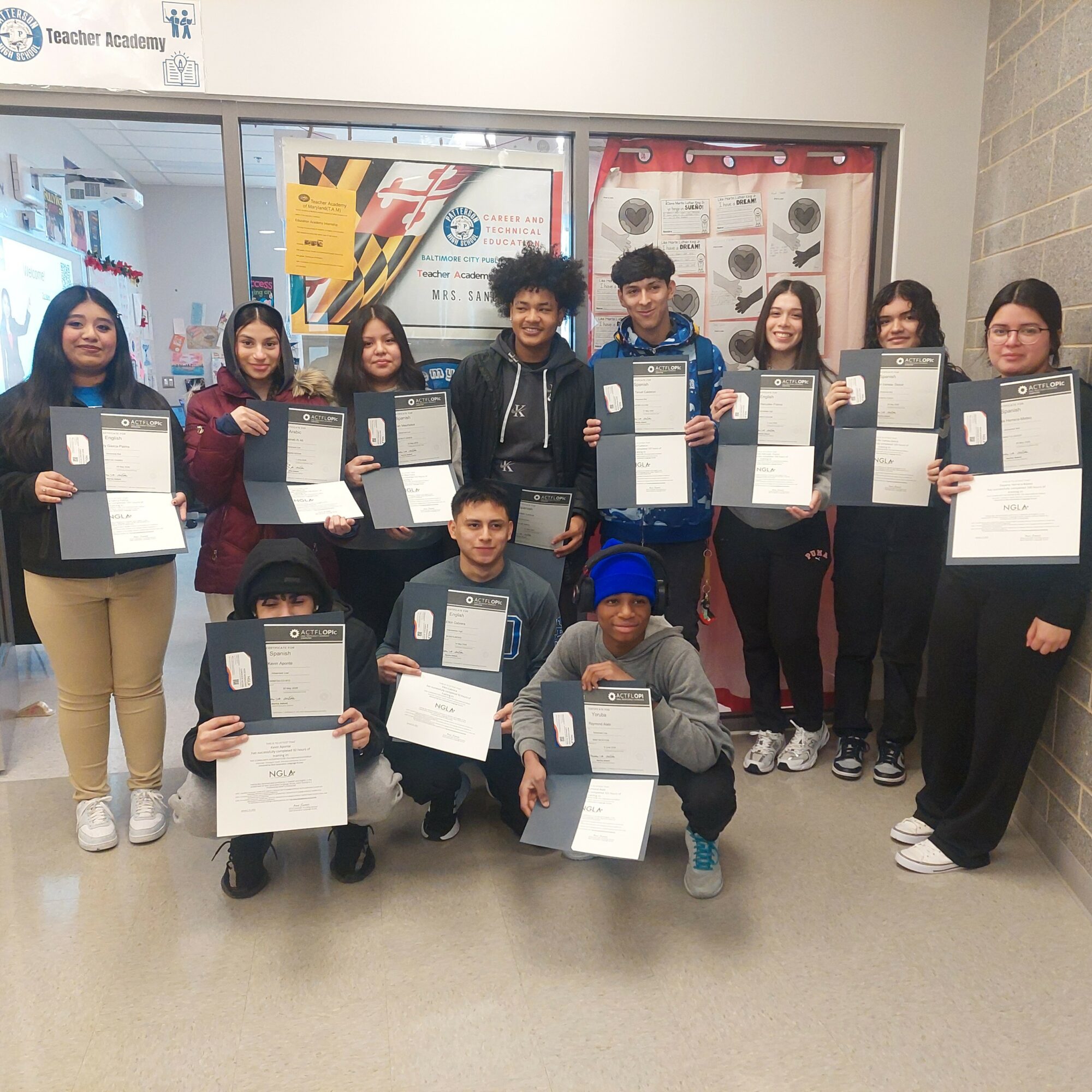 Group of ten young people holding certificates indoors, smiling, standing in front of glass doors with posters.