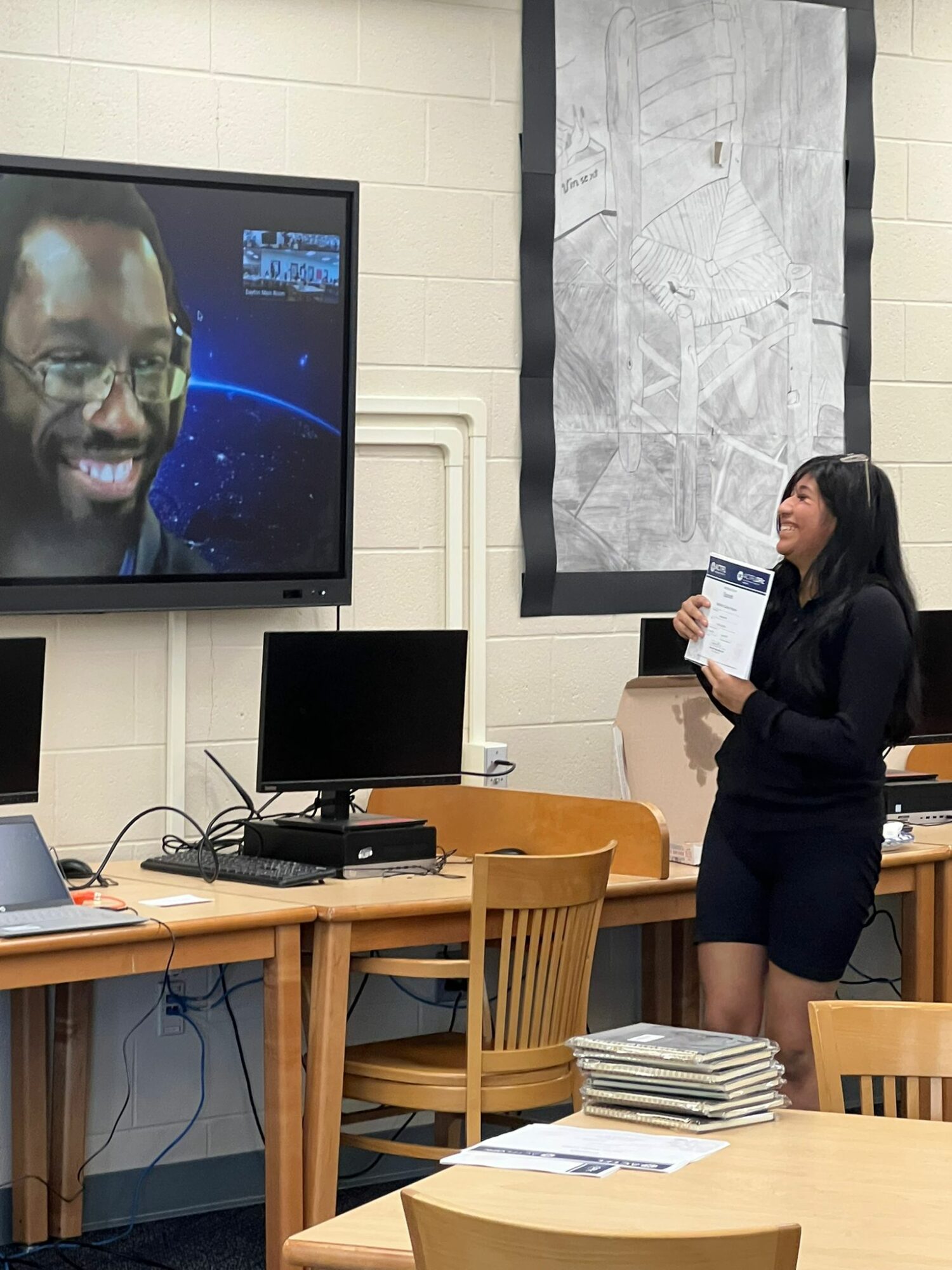 Woman standing in a classroom, holding a tablet, looking at a large screen with a man's face on it, in a classroom setting.