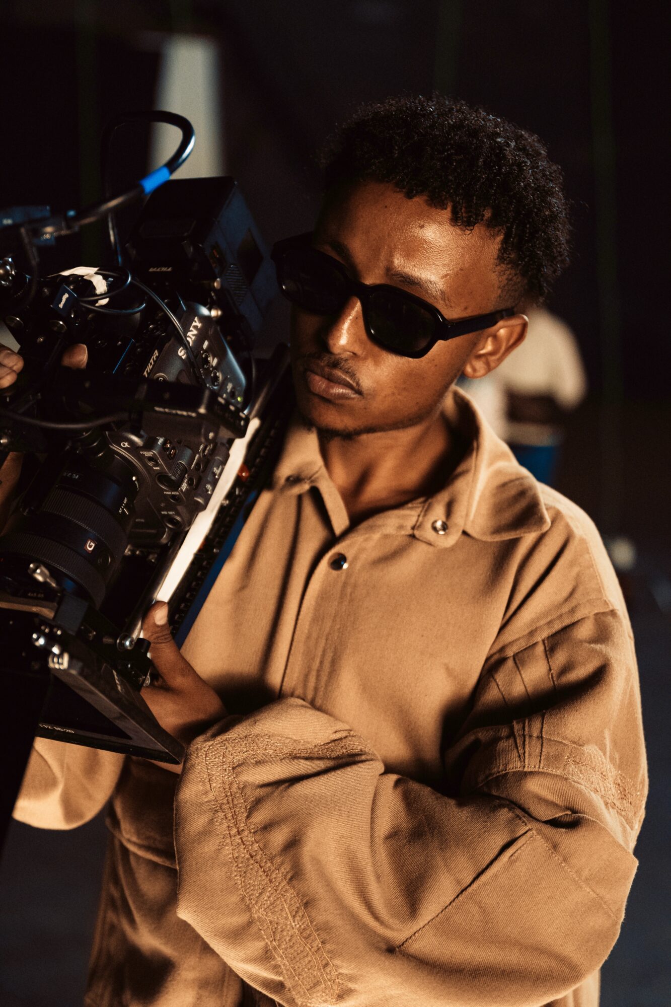 Person with sunglasses holding camera, wearing beige jacket, dark background.