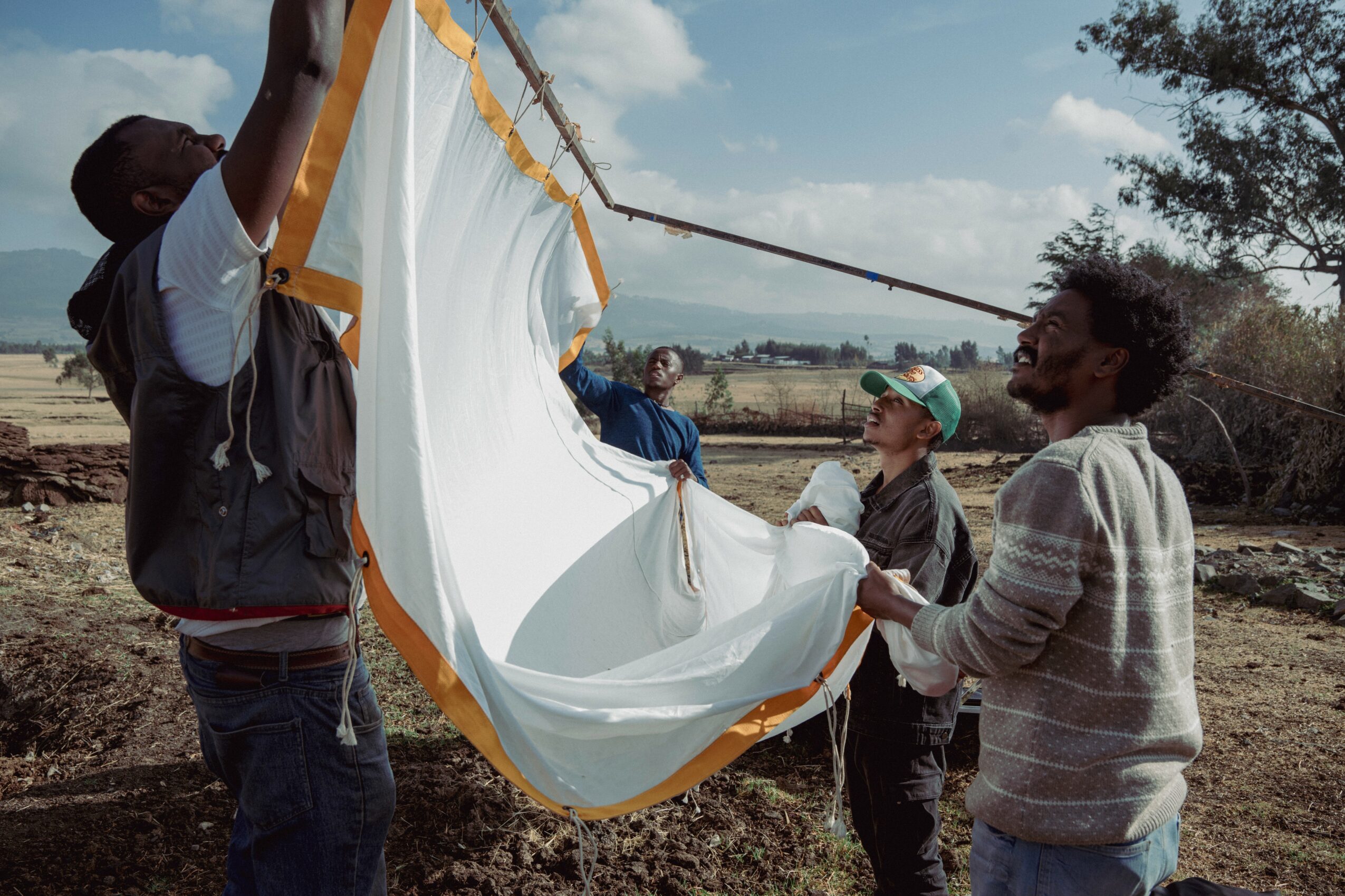 Four people outdoors holding a large white fabric with yellow trim, preparing to lift or unfold it.