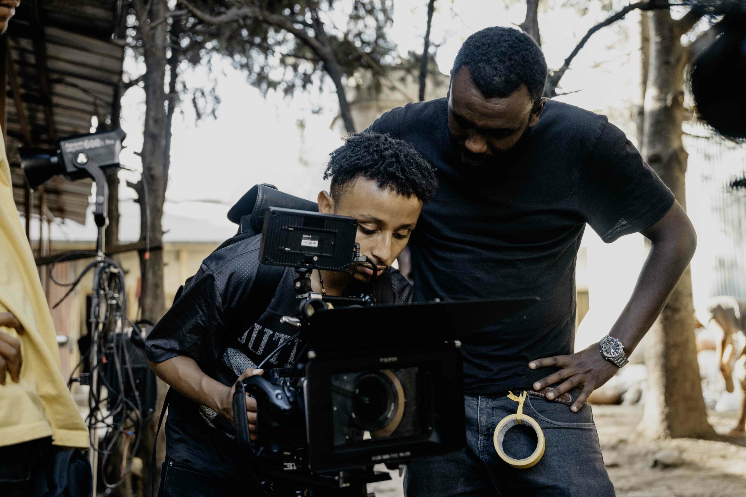 Two men outdoors, one operating a professional camera, the other observing, with trees and a fence in the background.