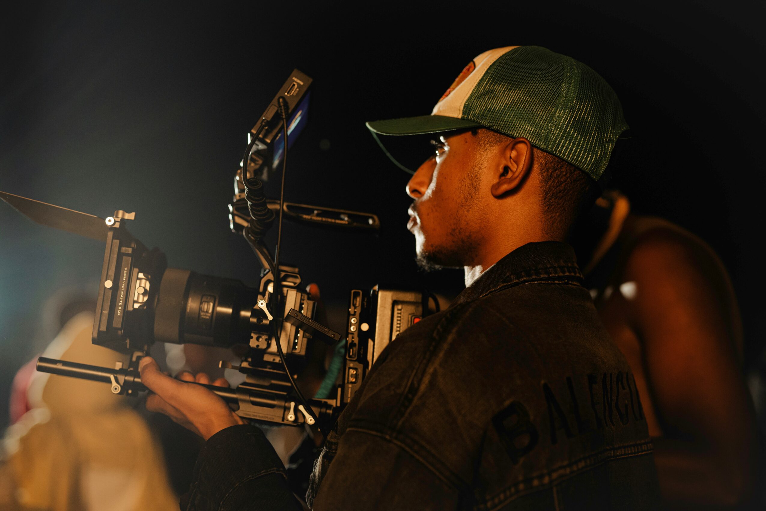Person operating a professional camera with a mounted microphone, wearing glasses and a cap, in a dark setting.