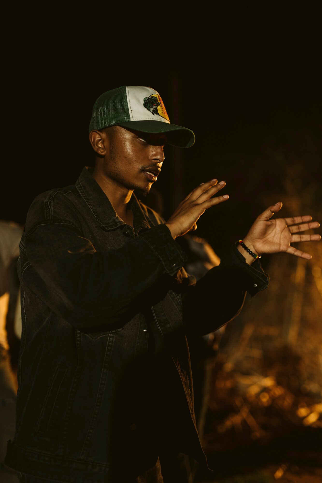 Person wearing a cap and black jacket, with hands raised, in a dark outdoor setting.