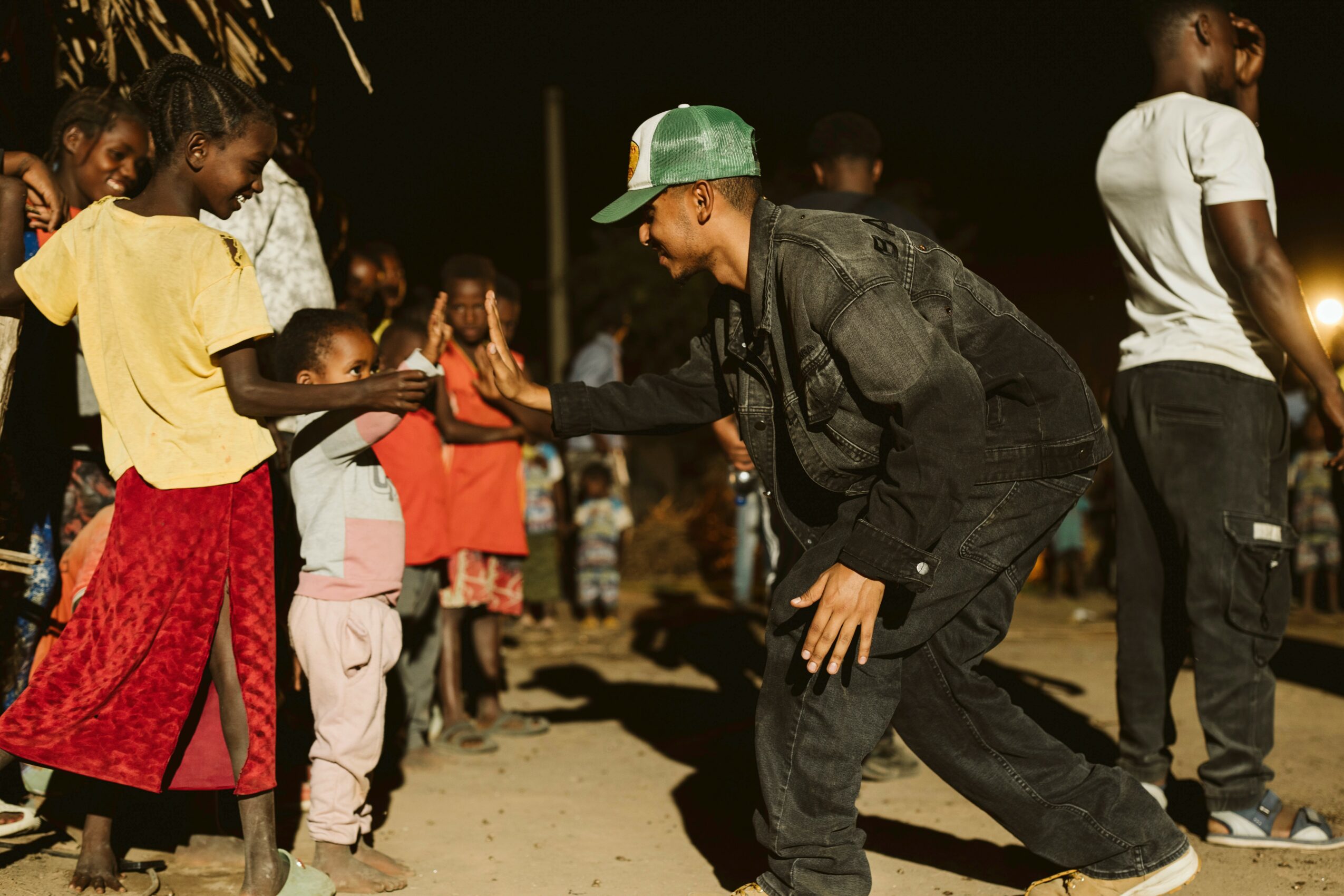 Person in a green cap and black jacket interacts with children outdoors at night, others standing nearby.