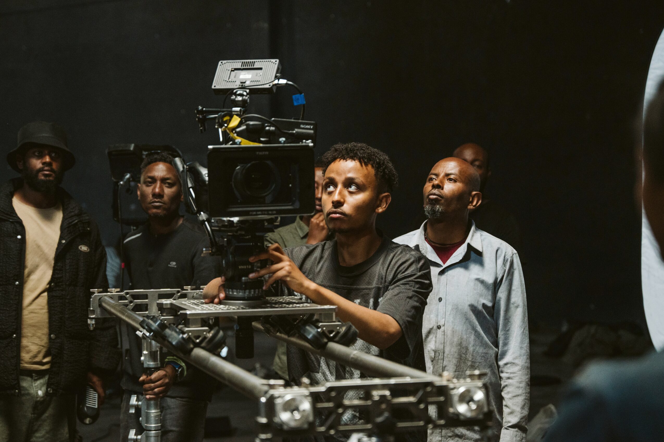 Group of five men on a film set, one operating camera equipment, others observing, dark background, professional environment.