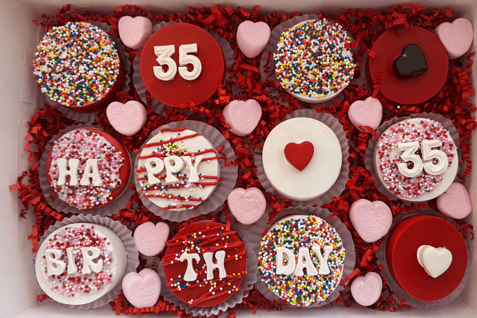 Assorted cupcakes with colorful sprinkles, hearts, and numbers celebrating a 35th birthday, decorated with red, pink, and white themes.