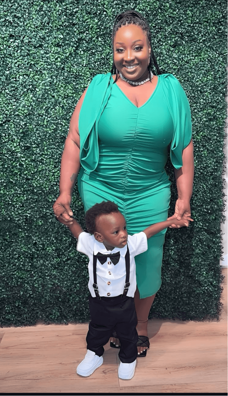 Woman in teal dress holding child's hands, standing in front of green hedge wall.