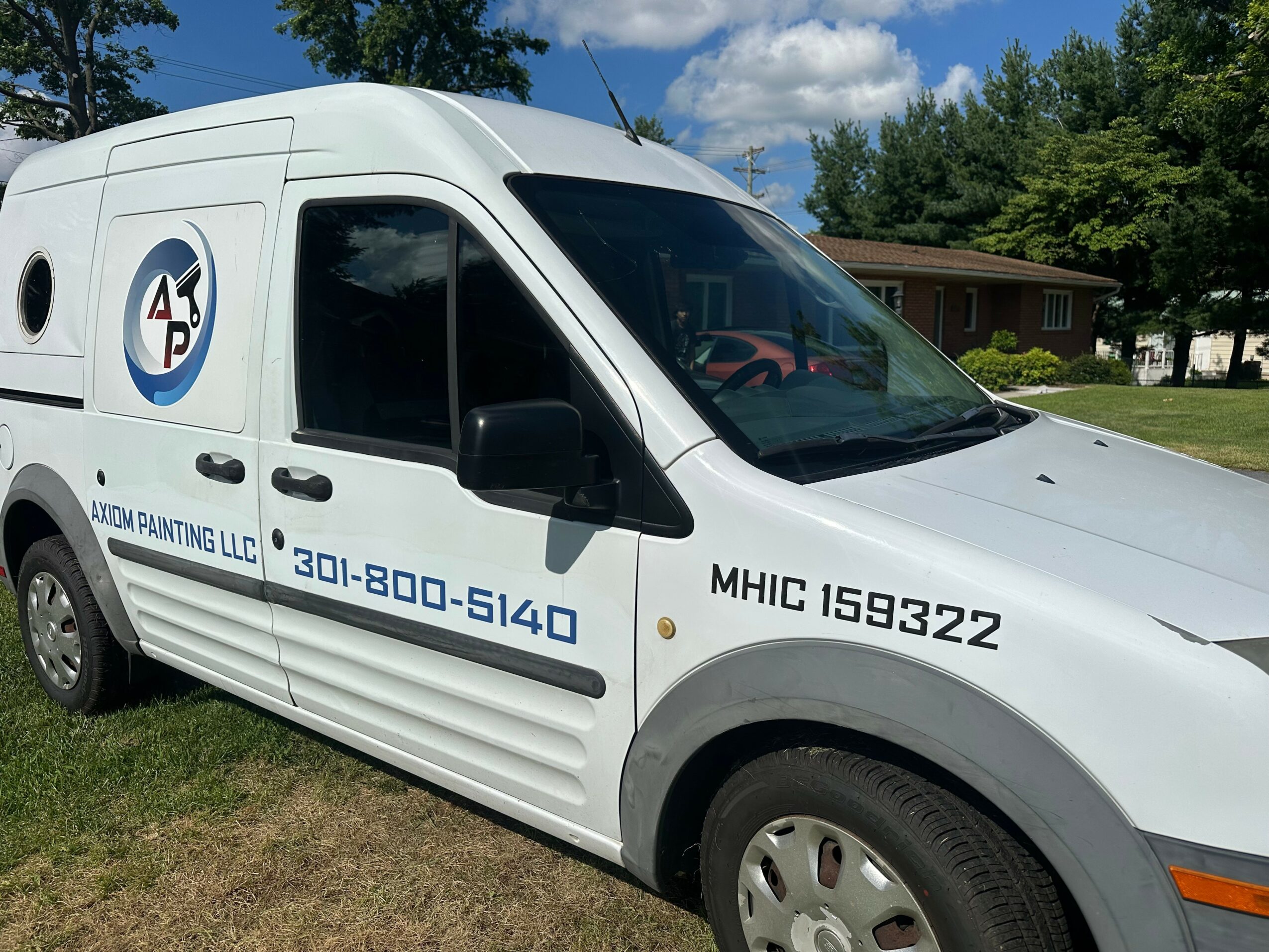 White van parked on grass with logo and contact number, house and trees in background, partly cloudy sky.
