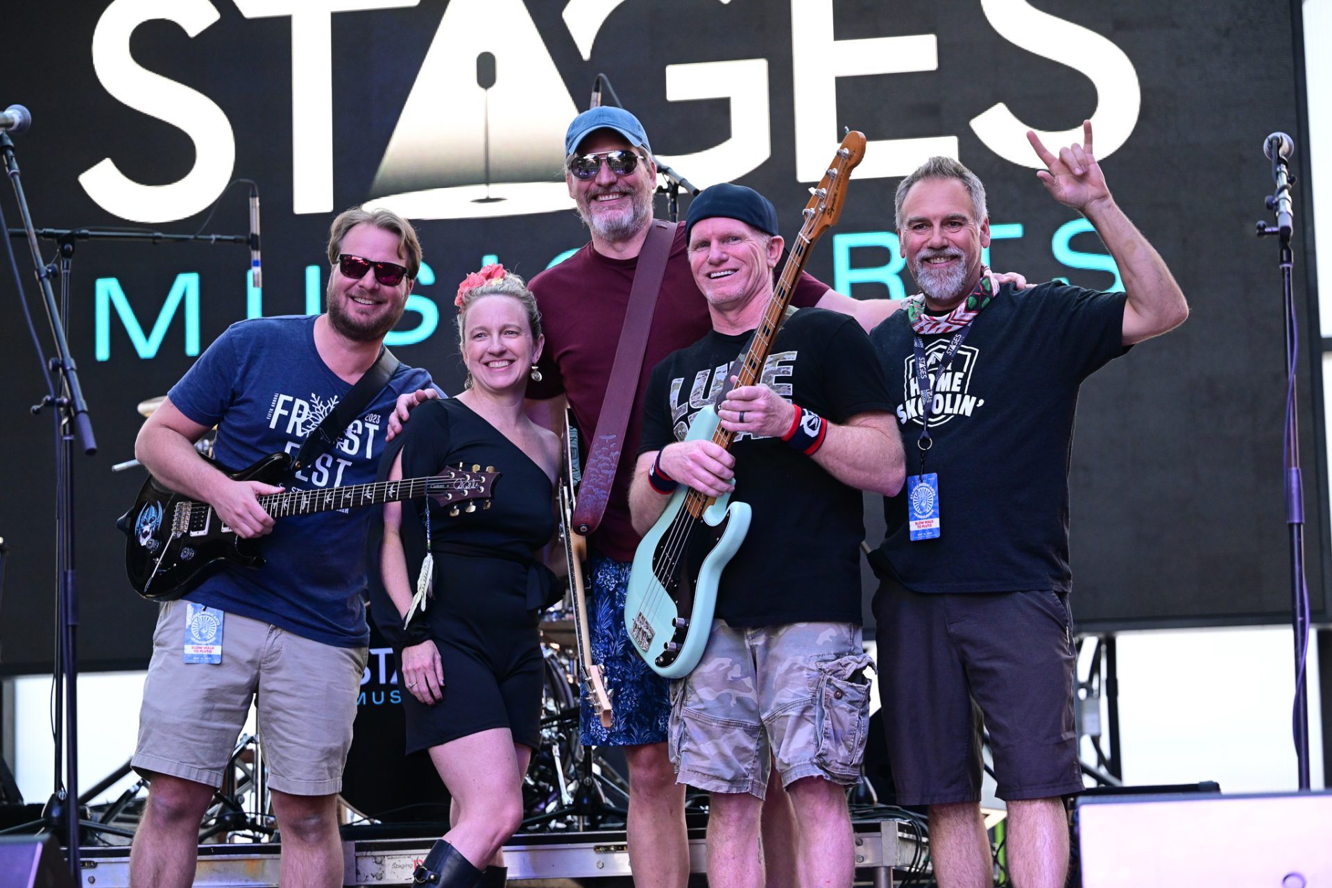 Group of five people on stage with musical instruments, smiling, in front of a large screen with text.