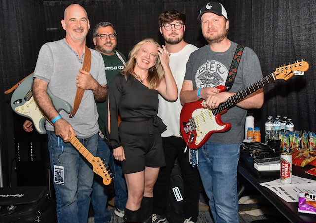 Group of five people, four men and one woman, standing with musical instruments and equipment behind them.