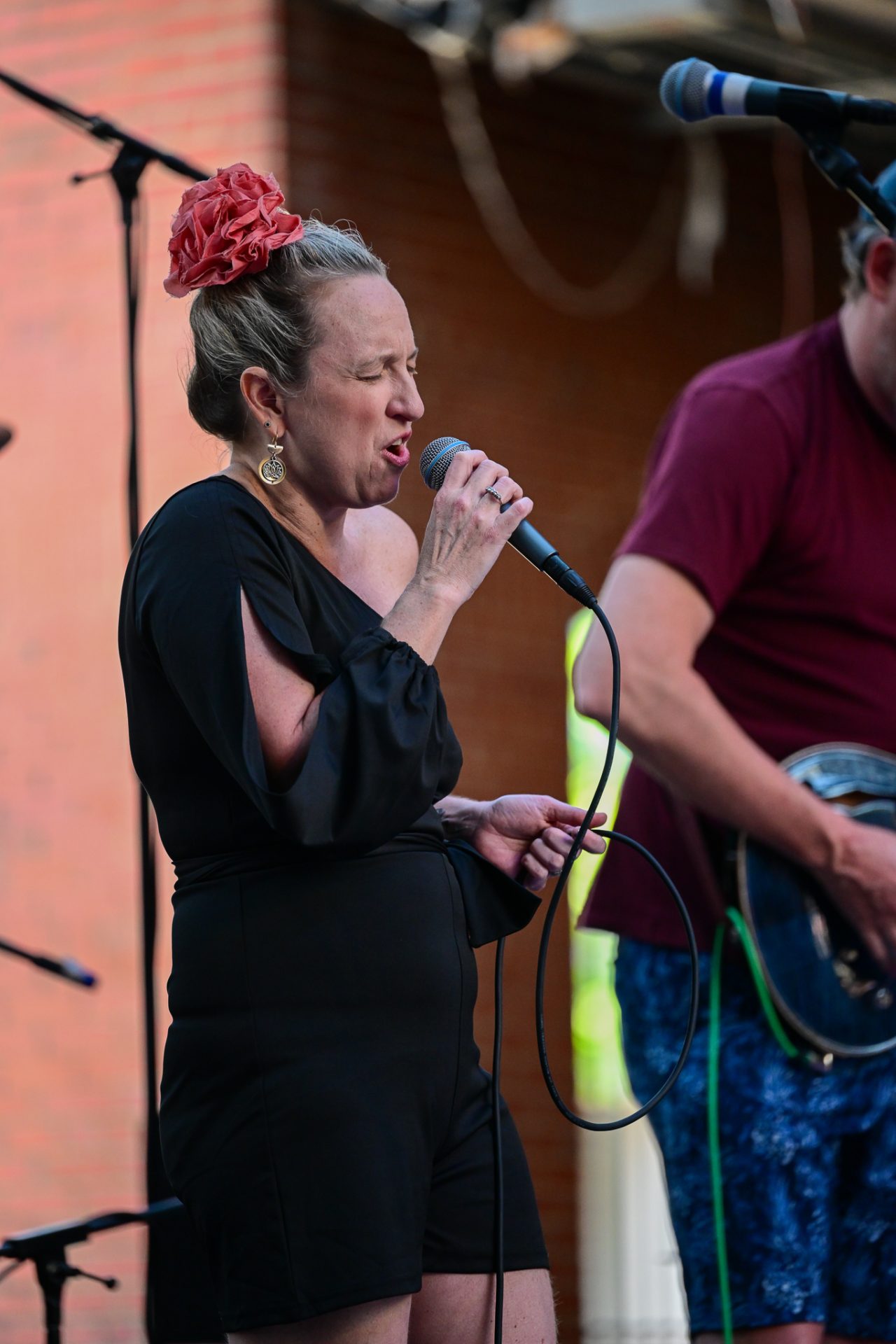 Woman singing into microphone on stage, wearing black outfit and red hair accessory, with a man playing guitar beside her.