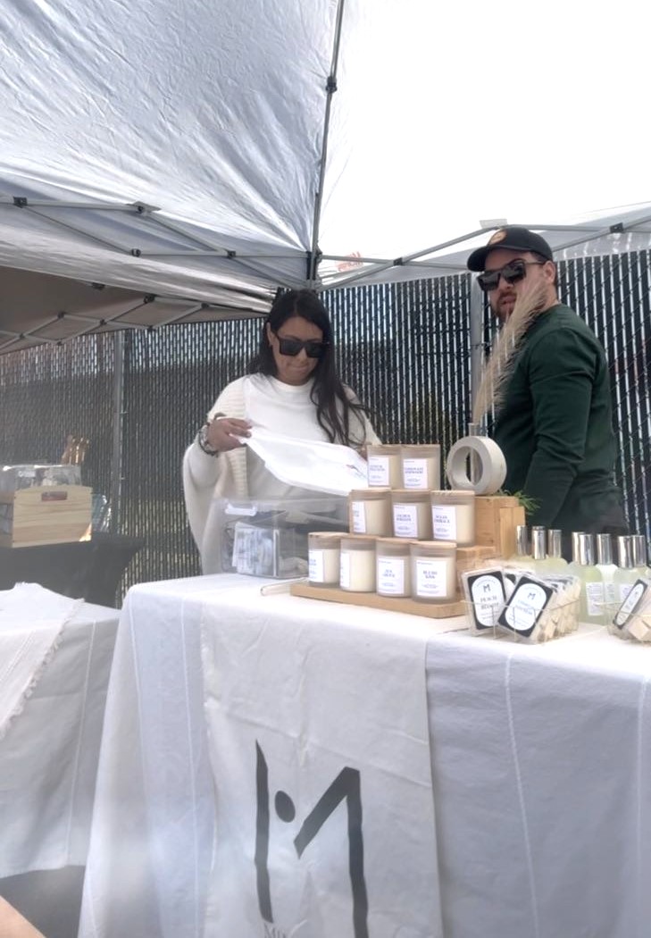Two women stand behind a table with jars and a scale, under a canopy outdoors, with a fence in background.