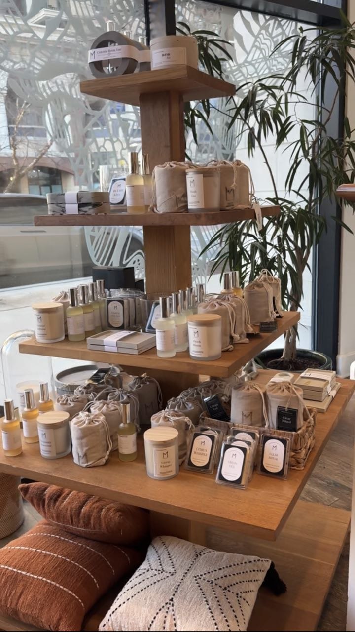 Wooden display stand with various skincare and cosmetic products in jars and bottles, set indoors near large windows and plants.