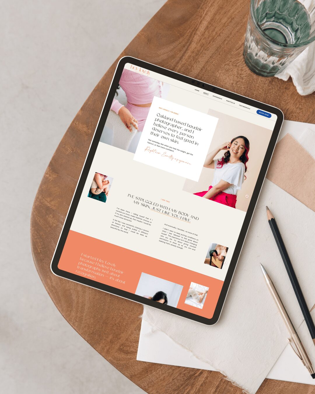 Tablet on wooden table displaying a website with images of a woman and text, next to a glass of water and a notebook.