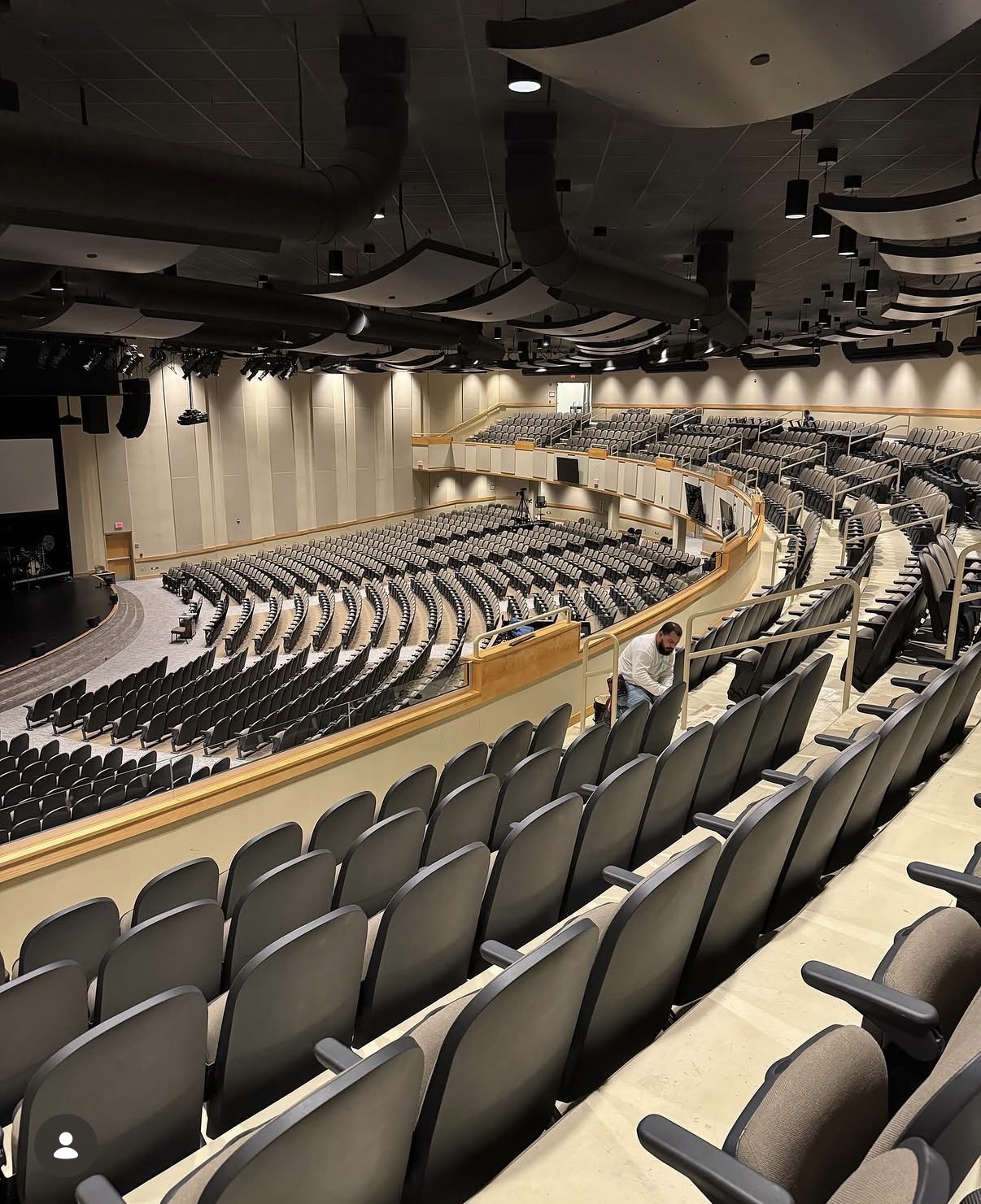 Empty auditorium with curved seating arrangement and stage at the front, ceiling with acoustic panels, and a person standing near seats.