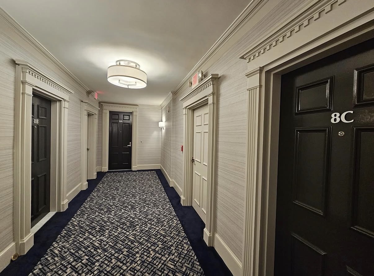 Hotel corridor with black doors, white walls, decorative molding, and patterned carpet, illuminated by ceiling light fixtures.