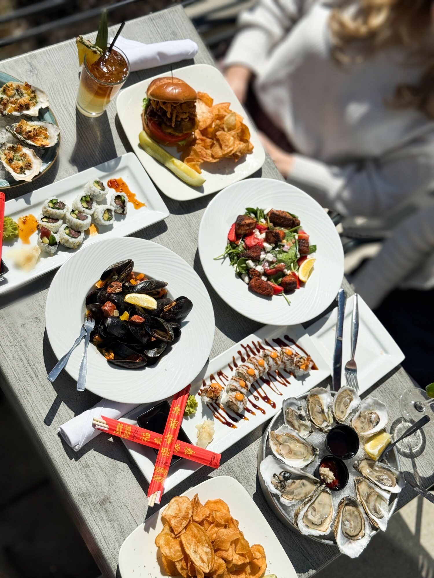 Table with various dishes including sushi, burger, salad, pasta, and fried chicken, with drinks and utensils, outdoors setting.