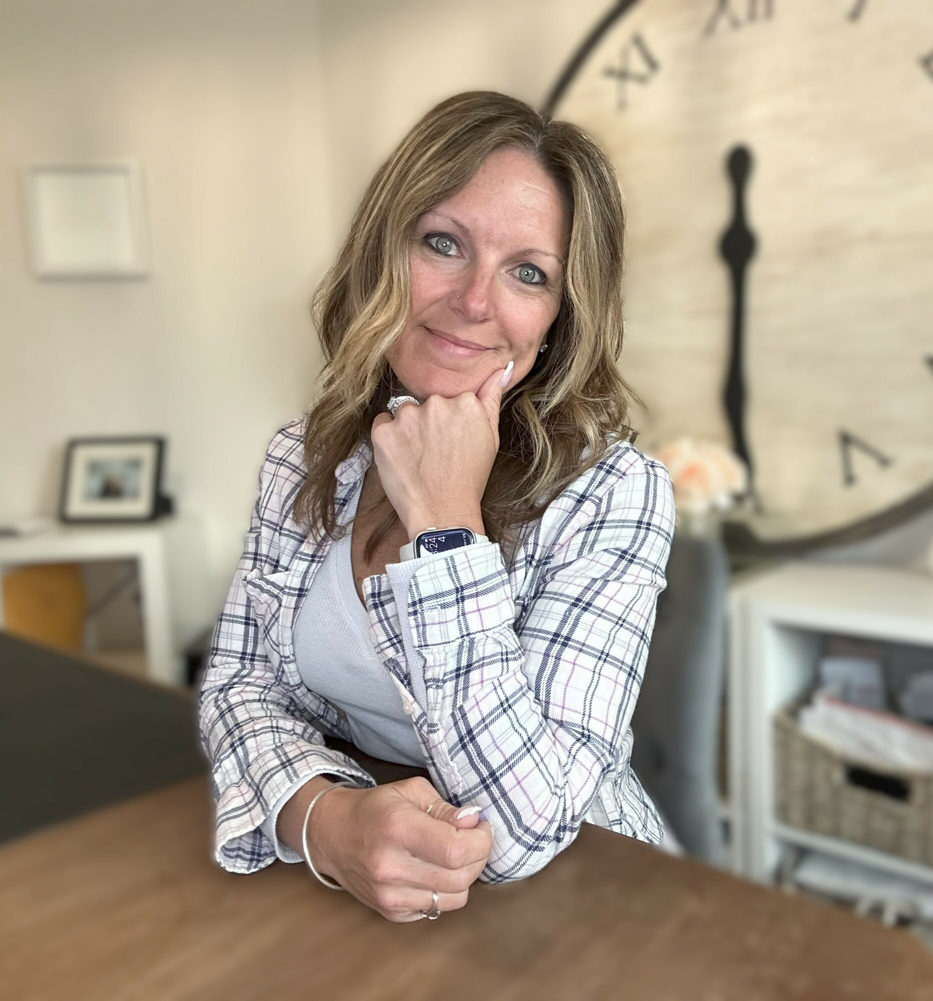 Woman with wavy hair smiling, resting chin on hand, in a room with clock and framed photo in background.