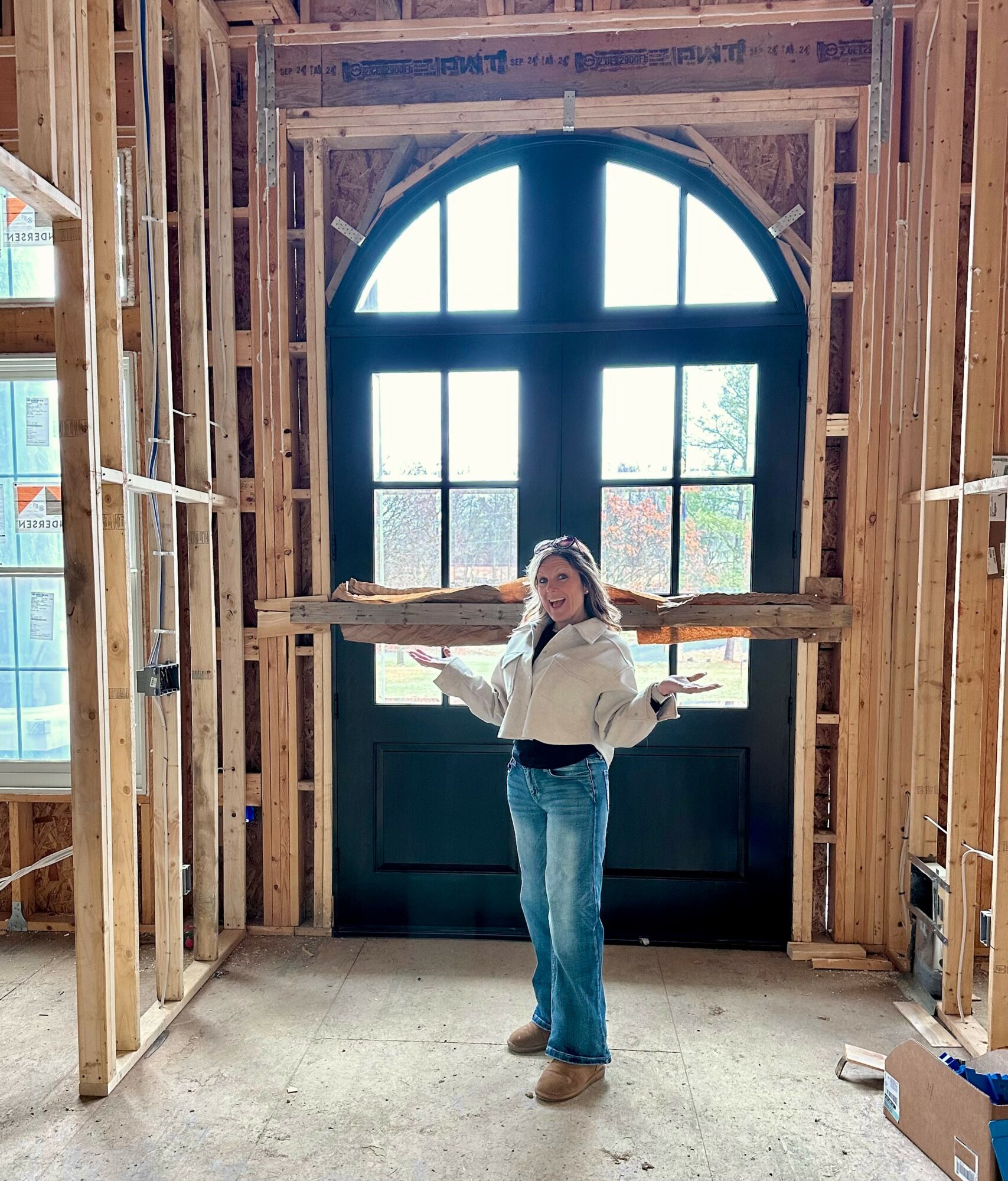 Person standing inside a wooden frame structure near a large arched window, holding a book or paper in each hand.