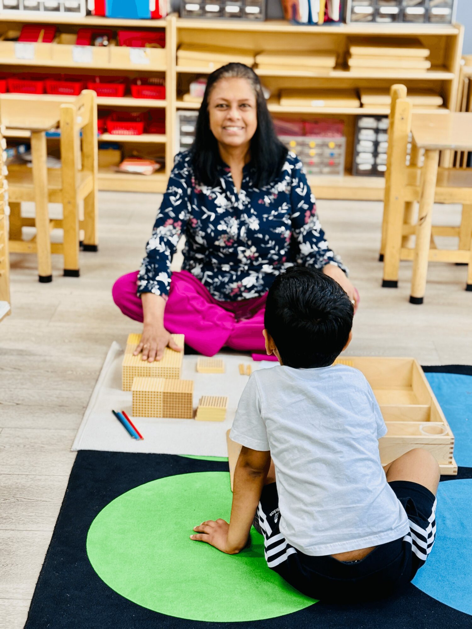 Woman sitting on the floor with a child, playing with blocks in a classroom setting.