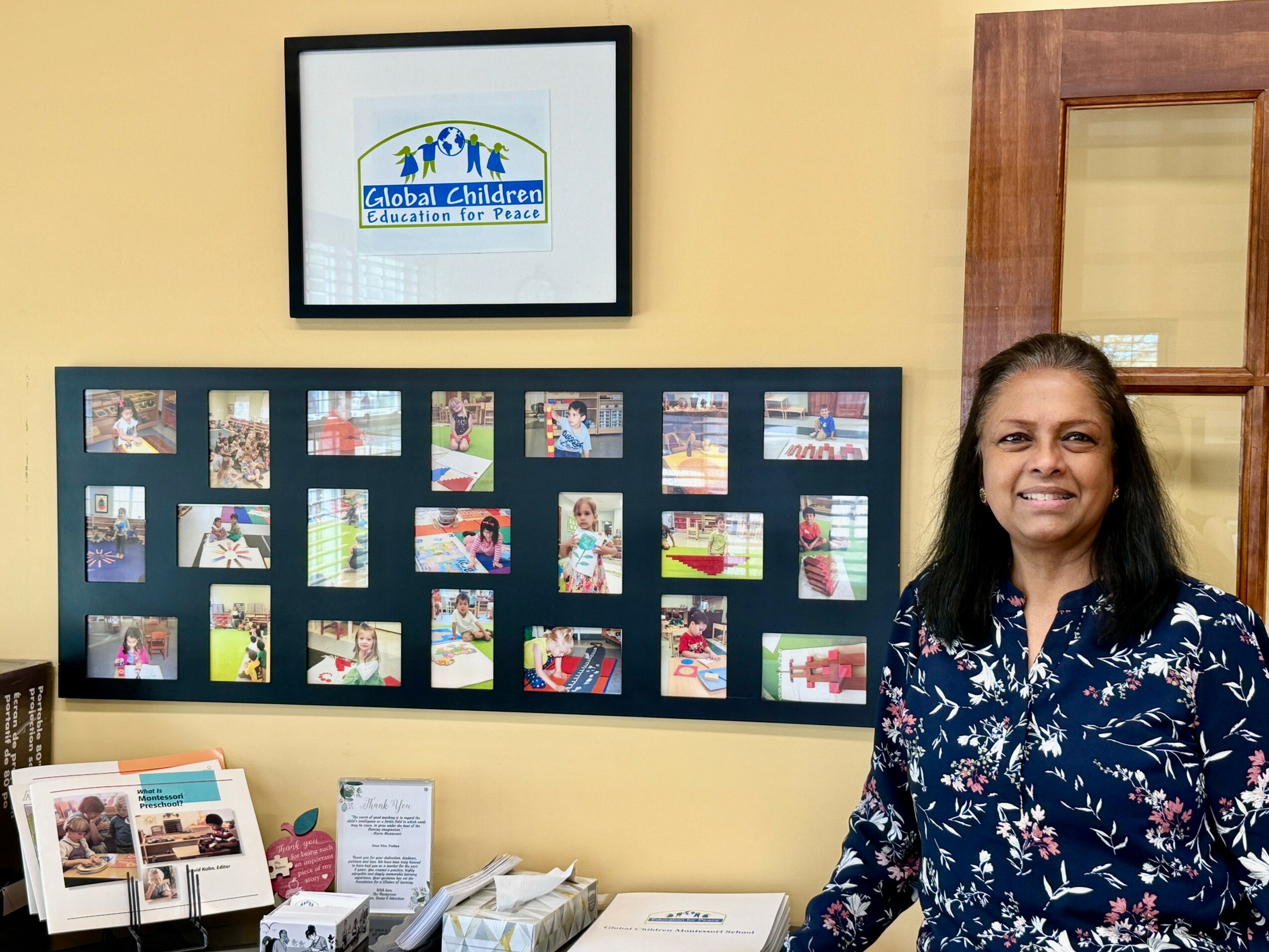 Woman with dark hair wearing a blue floral shirt standing indoors near a display board and framed screen.