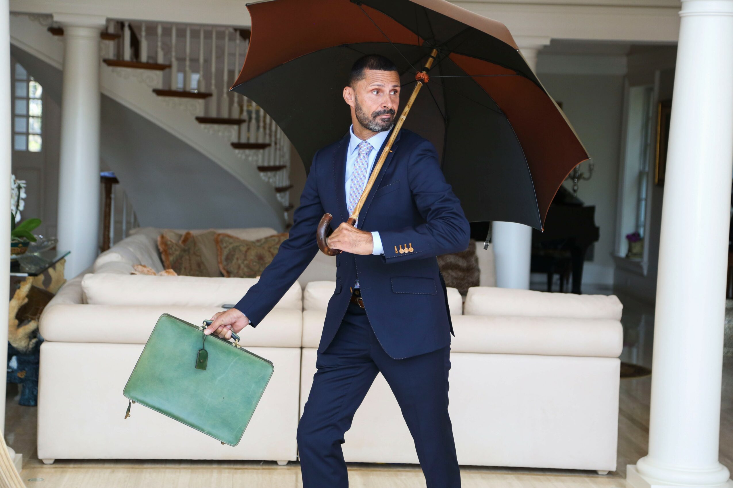 Man in a suit holding an umbrella and a briefcase in a living room with a sofa and staircase