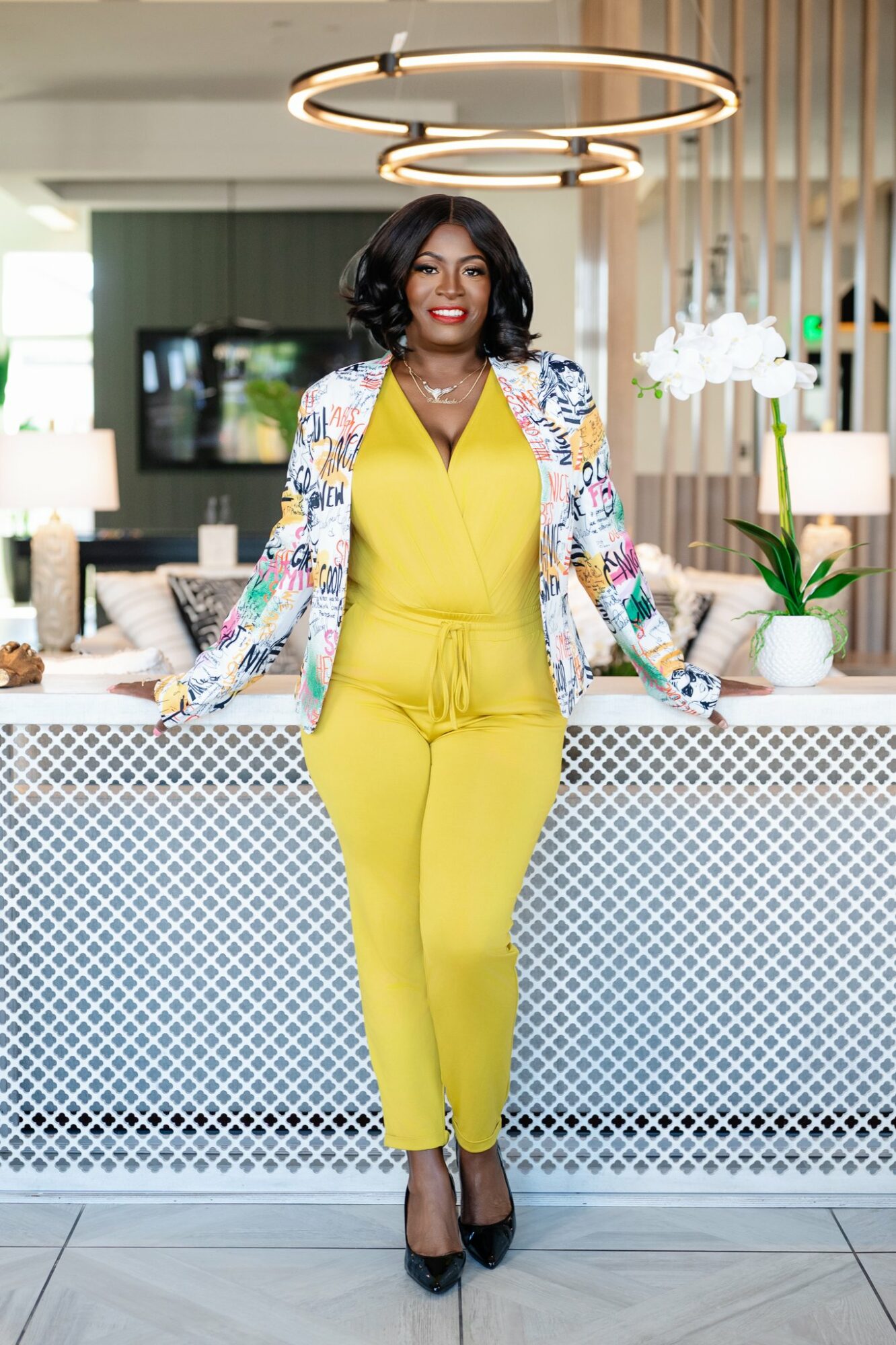 Woman in yellow jumpsuit and floral blazer standing in modern interior with circular ceiling light and white decor.