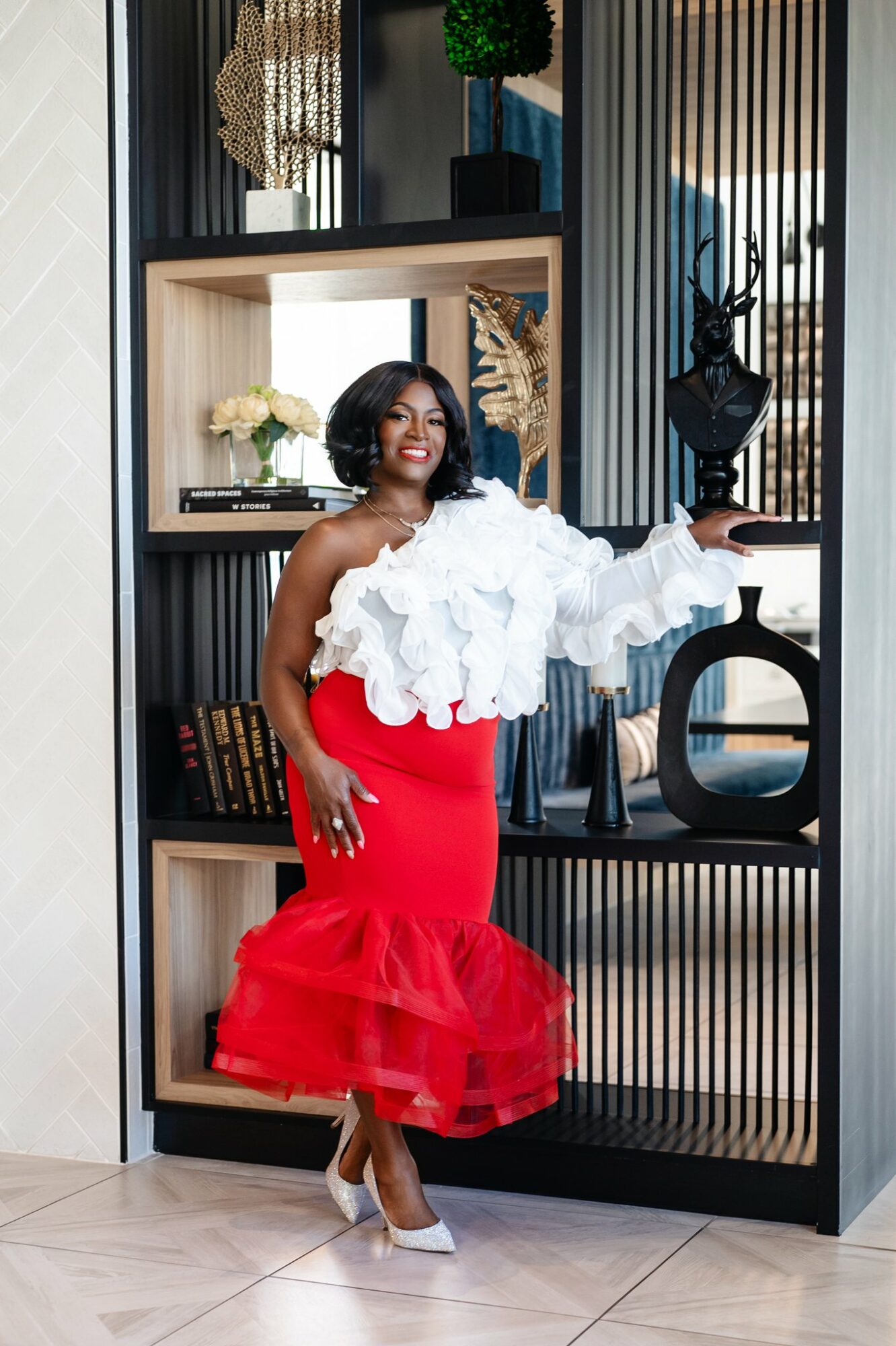 Woman in white ruffled top and red ruffled skirt standing next to a decorative shelf with plants and sculptures.