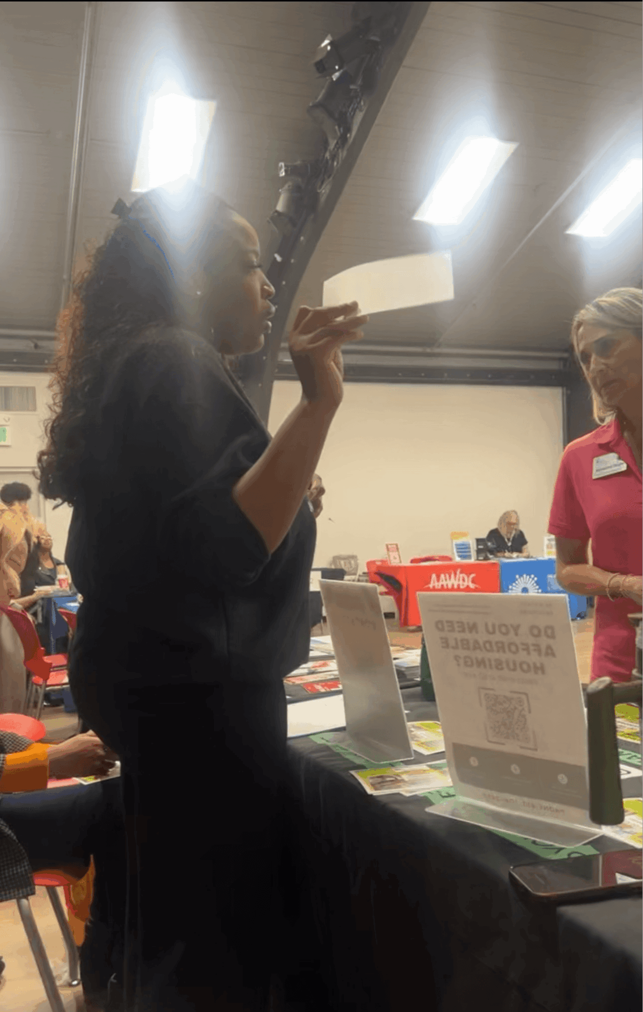 Person with curly hair holding a paper, standing at a table with informational signs, in an indoor setting.