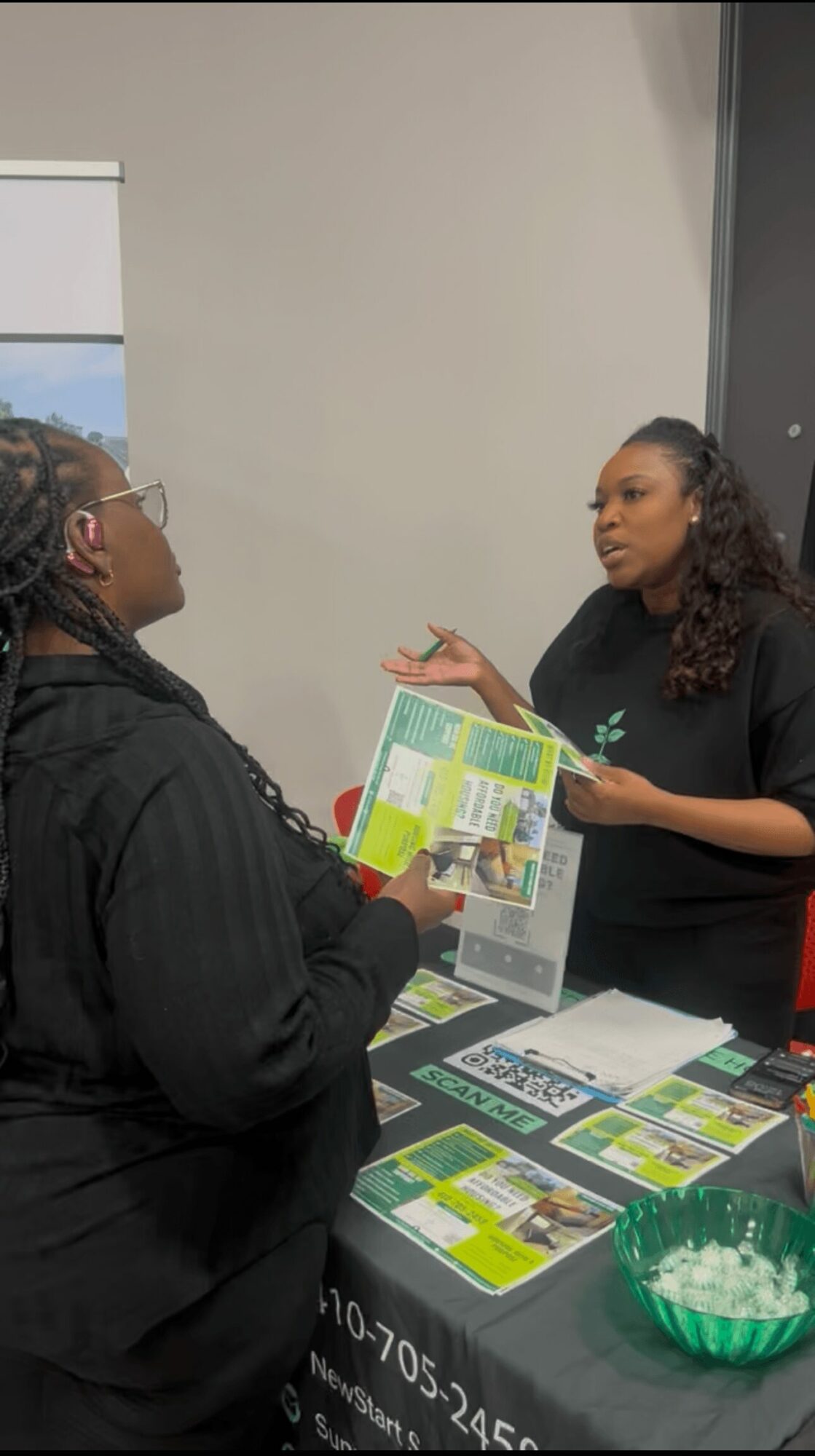 Two women engaged in conversation at a table with pamphlets and a bowl of green items.
