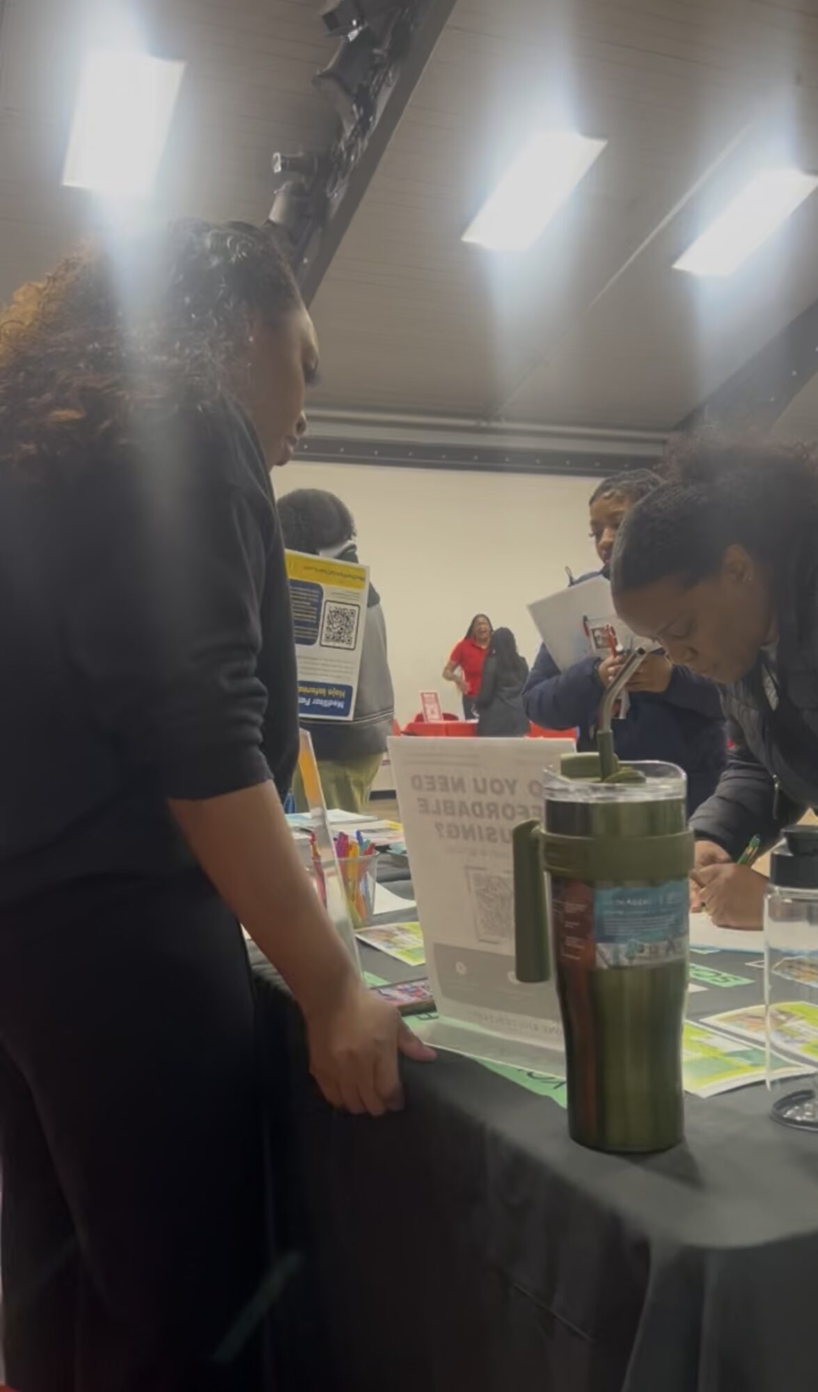 People gathered around a table with books and a green tumbler in a room with bright ceiling lights.