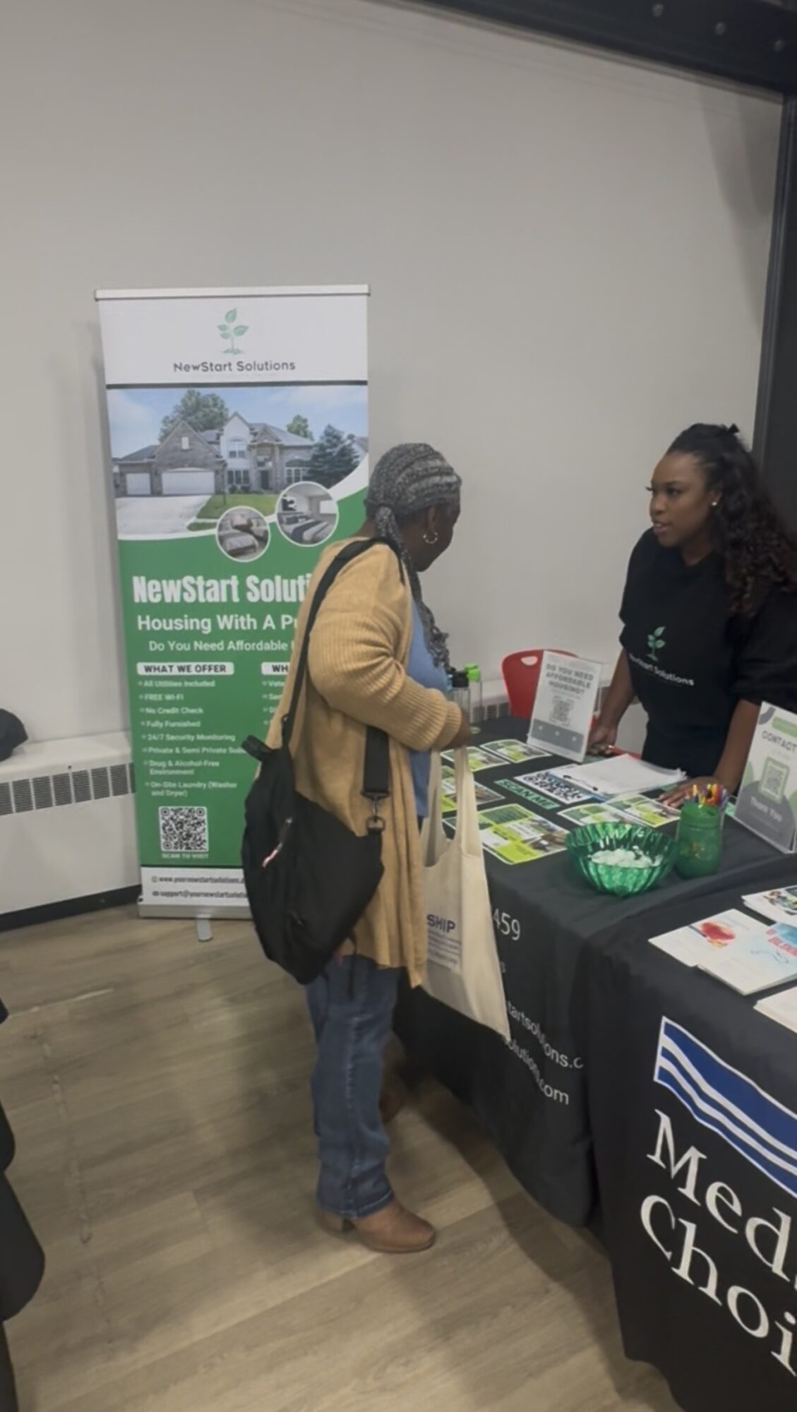 Two women talking at an informational booth with green and white materials, indoors with a plain wall background.
