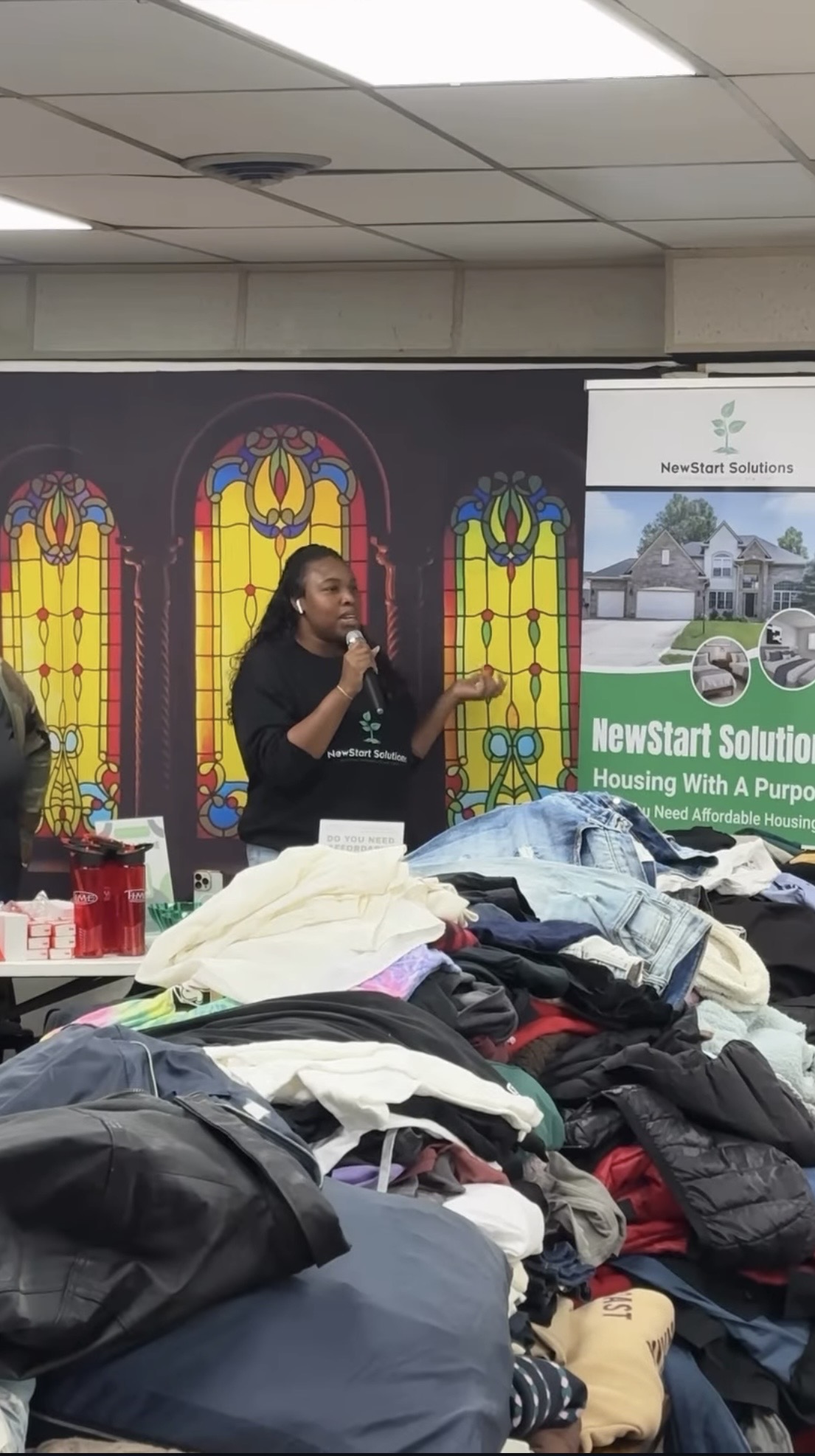 Woman speaking into microphone in front of stained glass windows, with a table of clothes and a banner behind her.