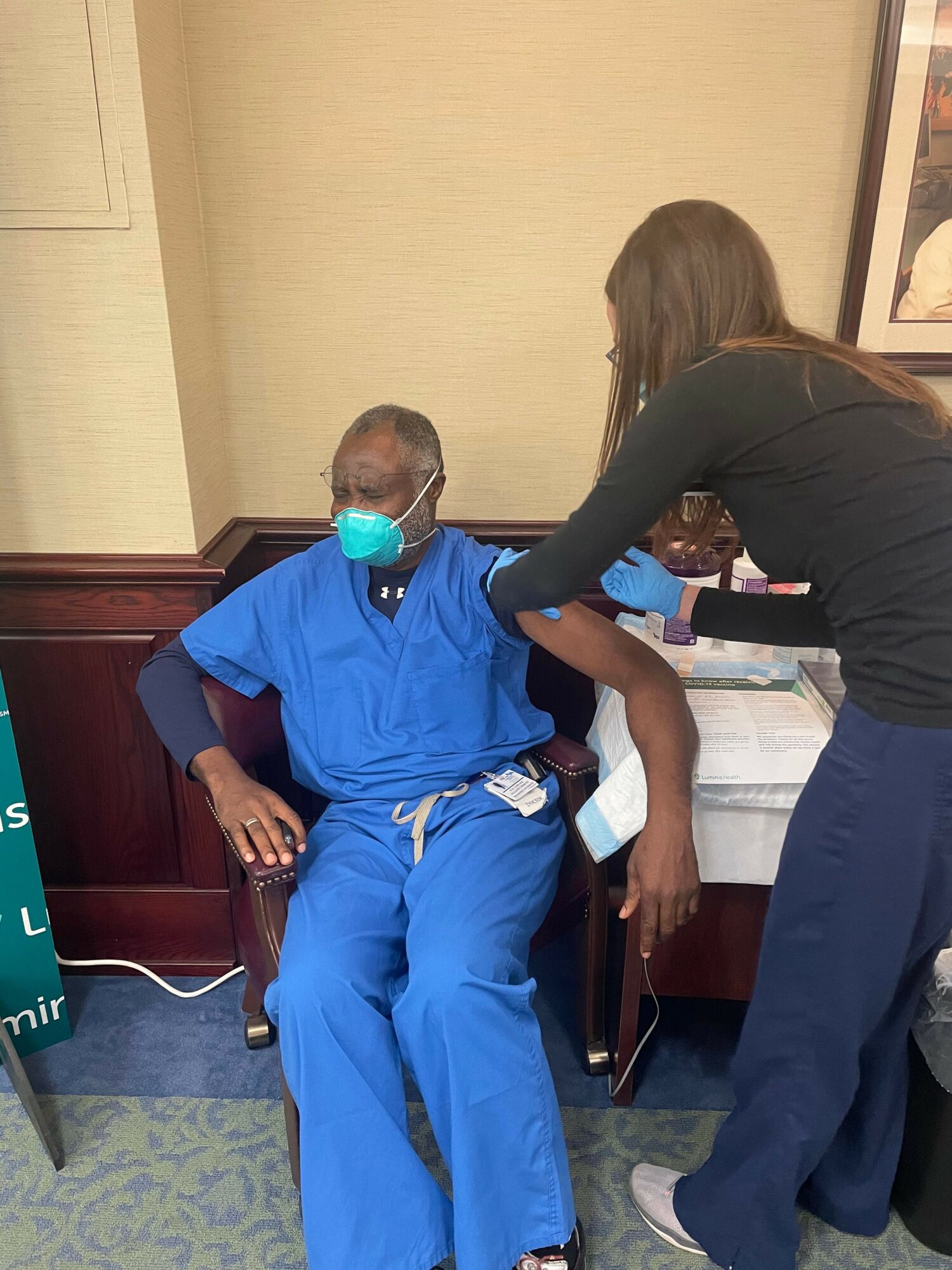 Healthcare worker in black uniform administers vaccine to seated person in blue scrubs, wearing a mask and glasses.