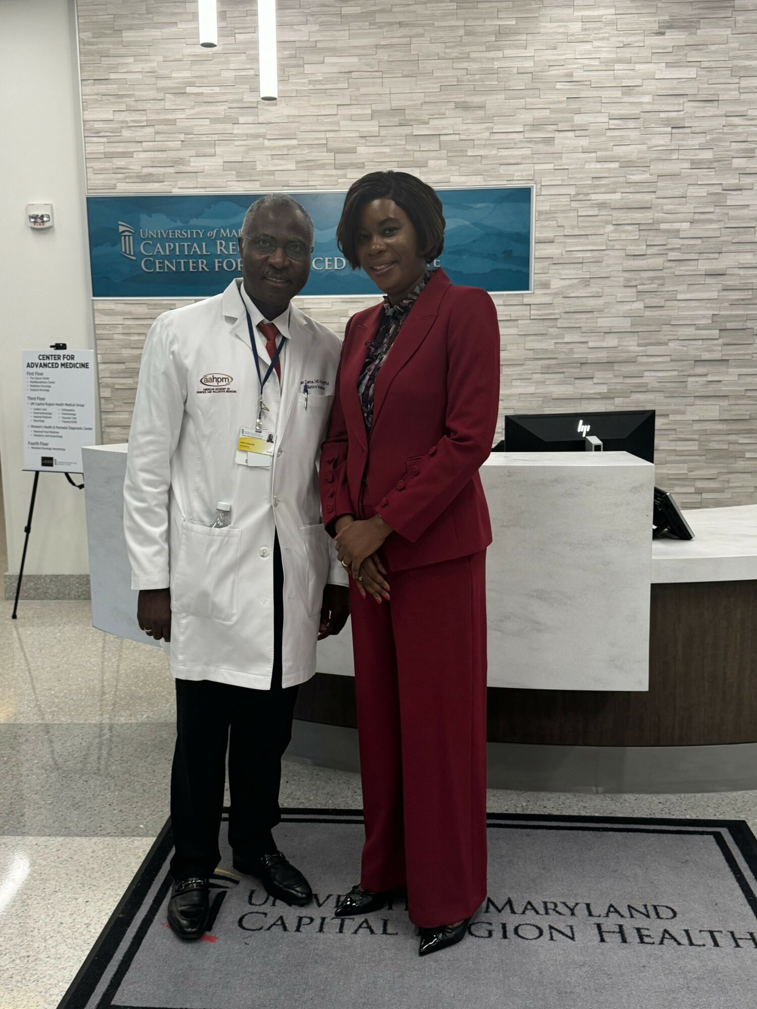 Two people, a man in a white coat and a woman in a red suit, stand together in a medical facility.