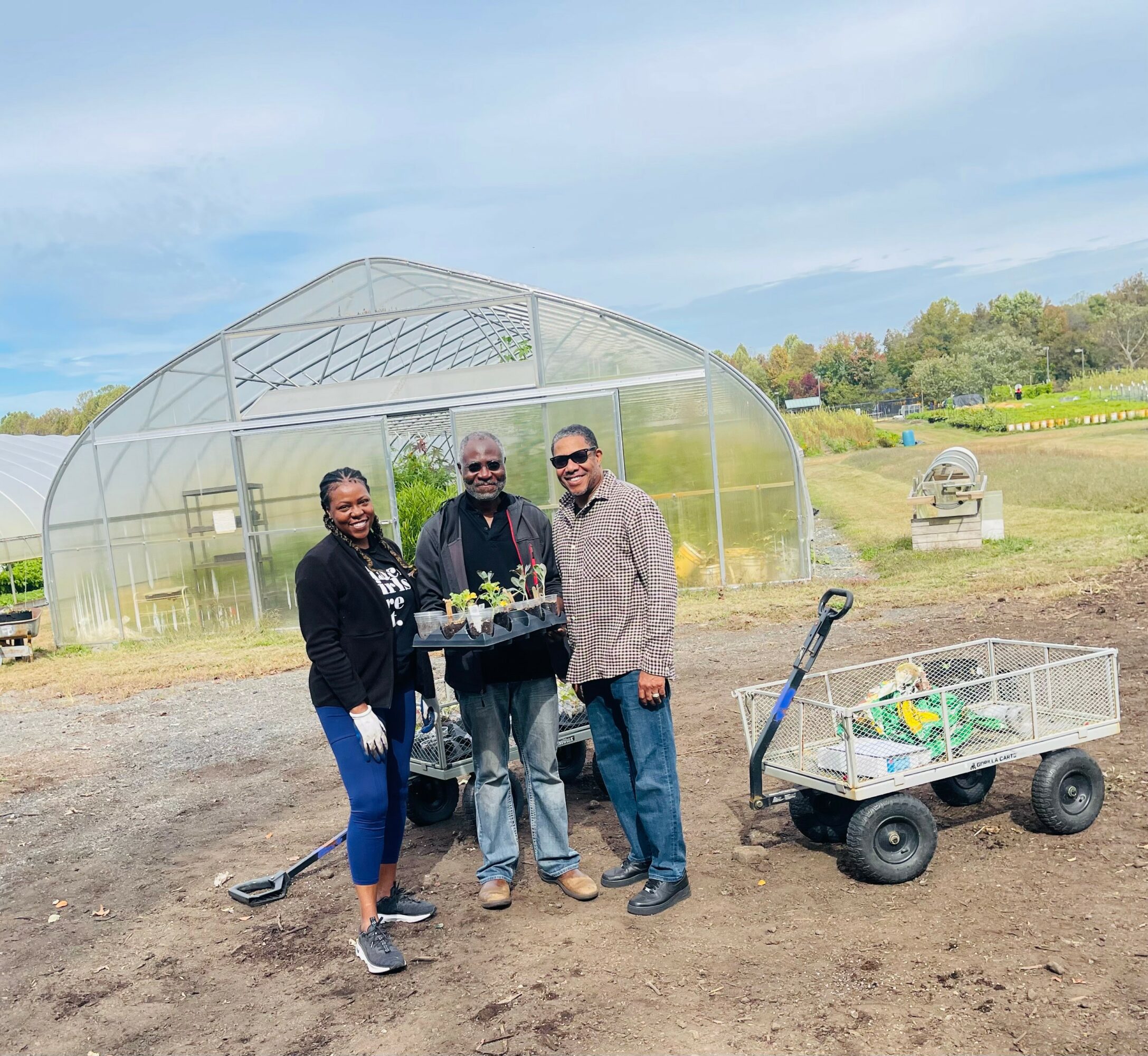 Three people standing outdoors near a greenhouse, holding a tray of plants, with a cart nearby and trees in the background.