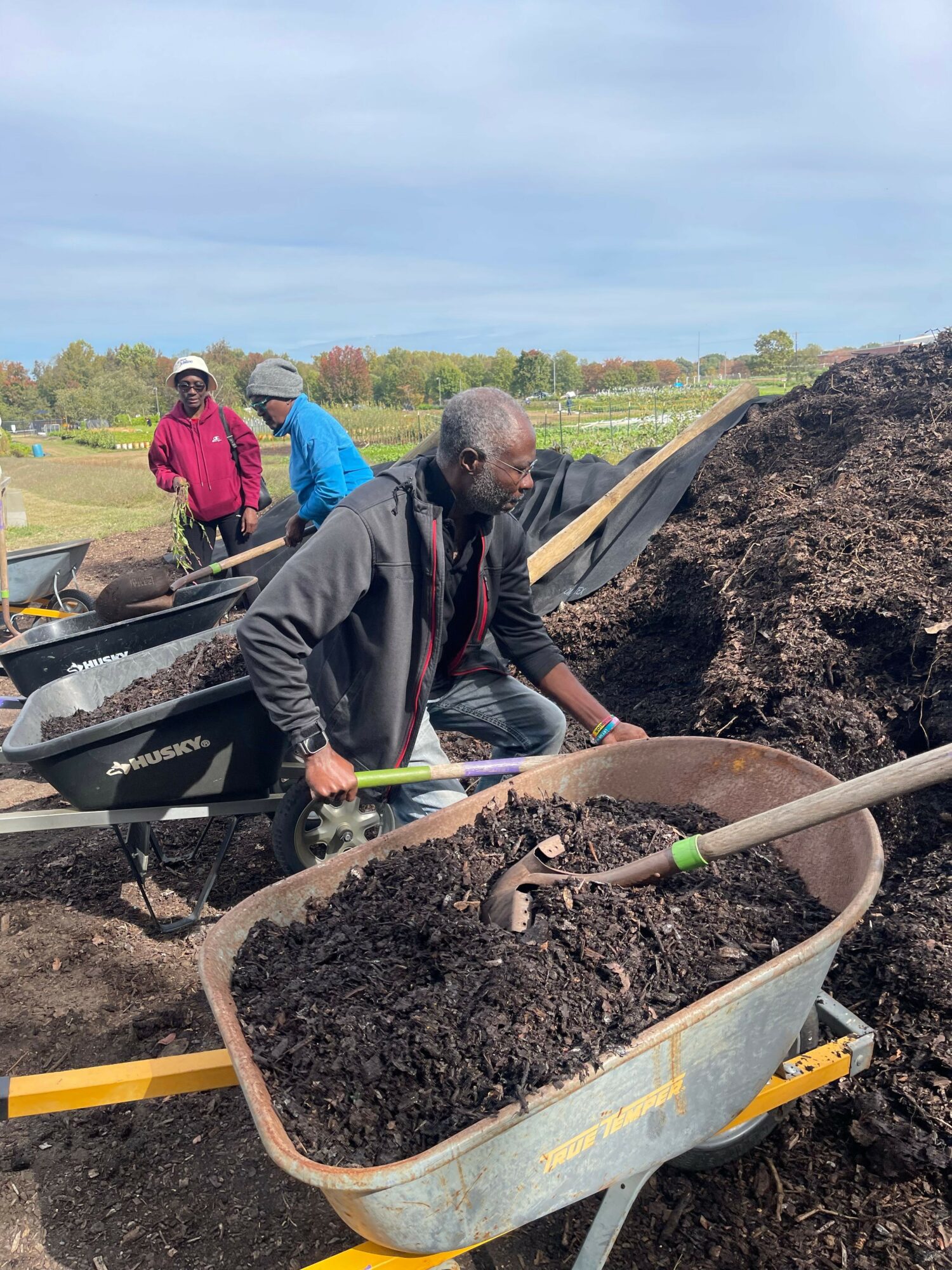 Man working with soil in a wheelbarrow outdoors, with two people in the background and a landscape with trees.