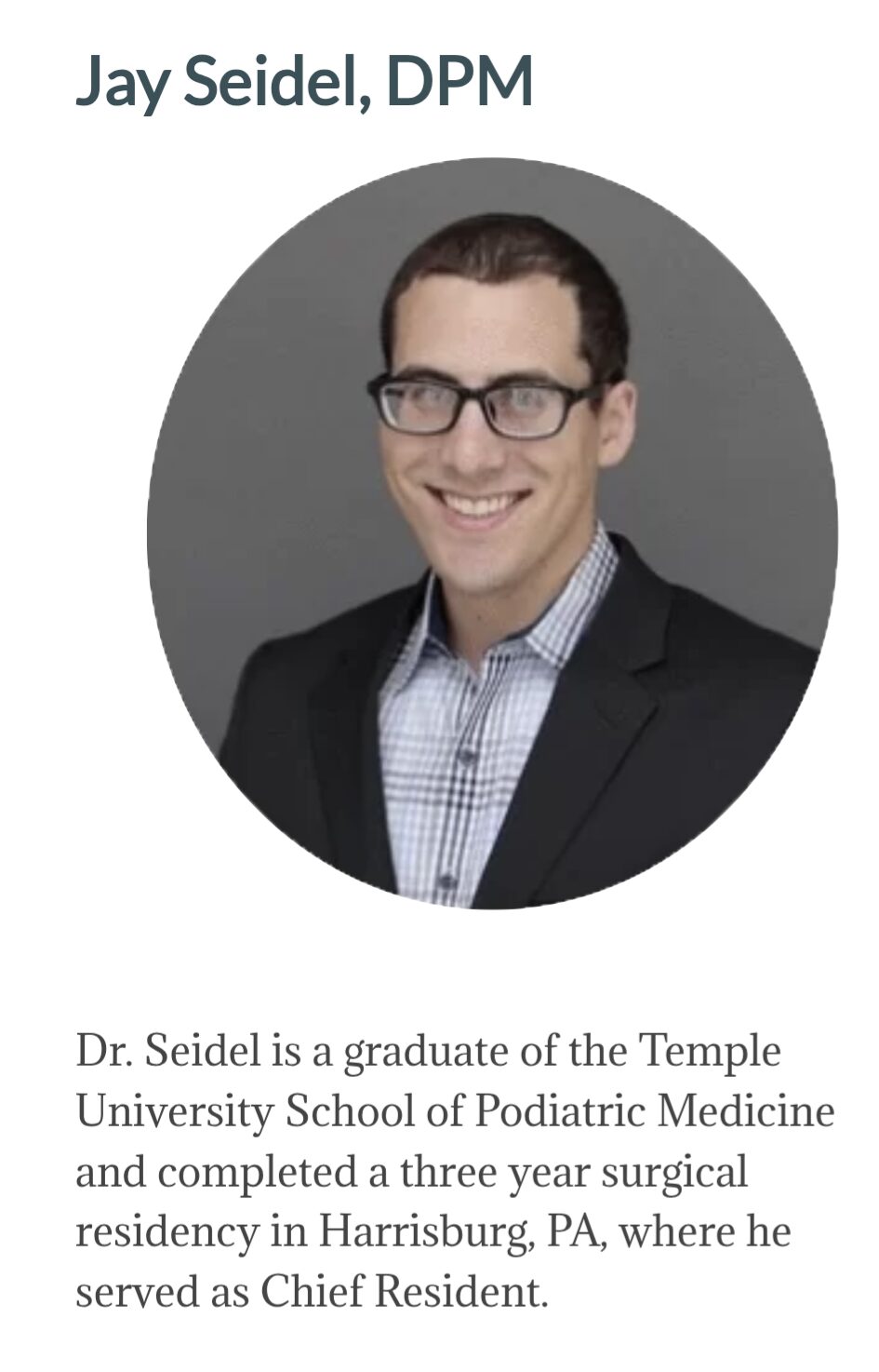 Professional portrait of a man with glasses, wearing a suit and checkered shirt, smiling against a gray background.