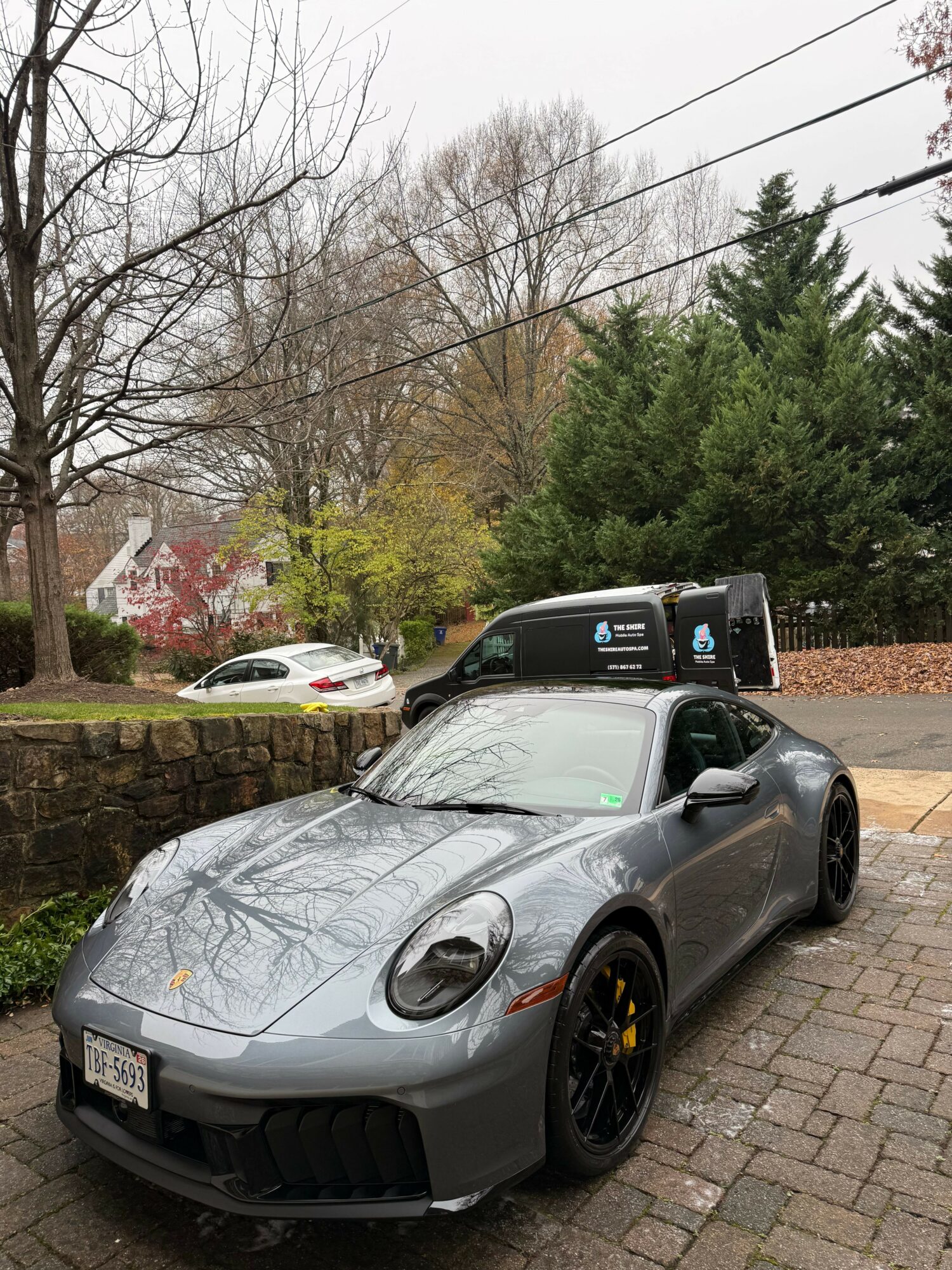 Gray sports car parked on driveway with trees and a house in background.