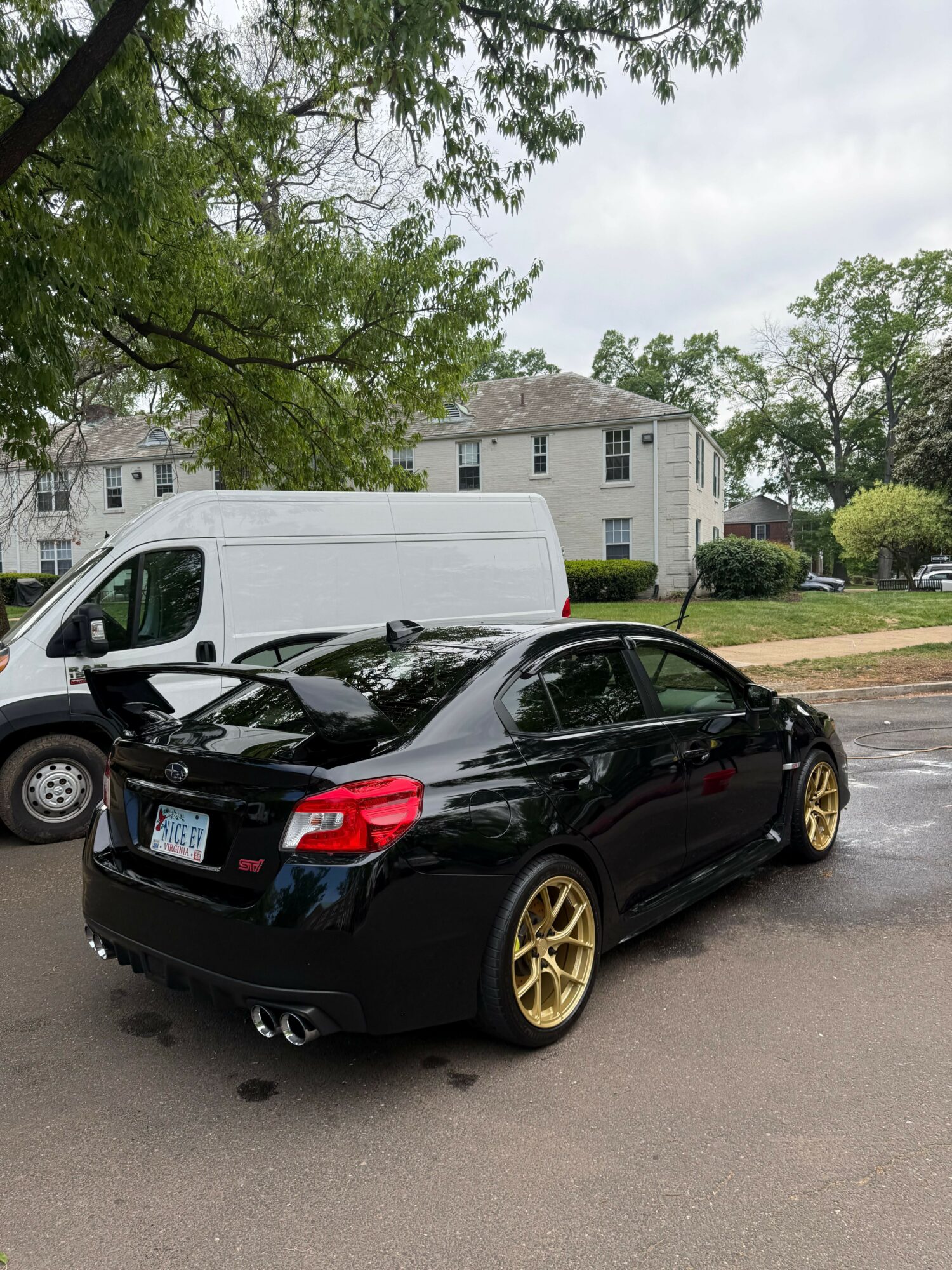 Black sports car with gold wheels parked on street, white van behind, residential houses and trees in background.
