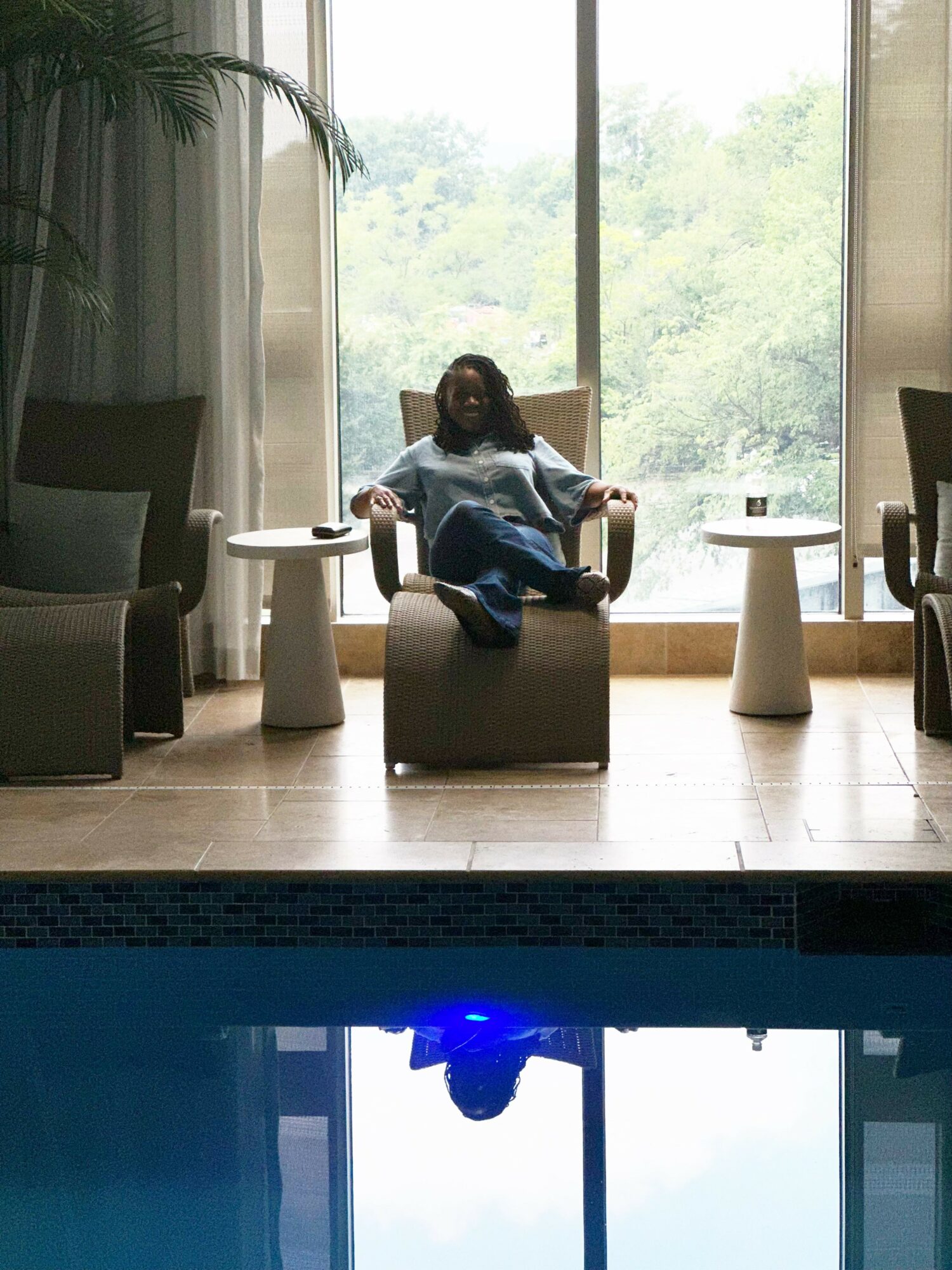 Person sitting in a chair by large window with trees outside, surrounded by small tables and chairs.