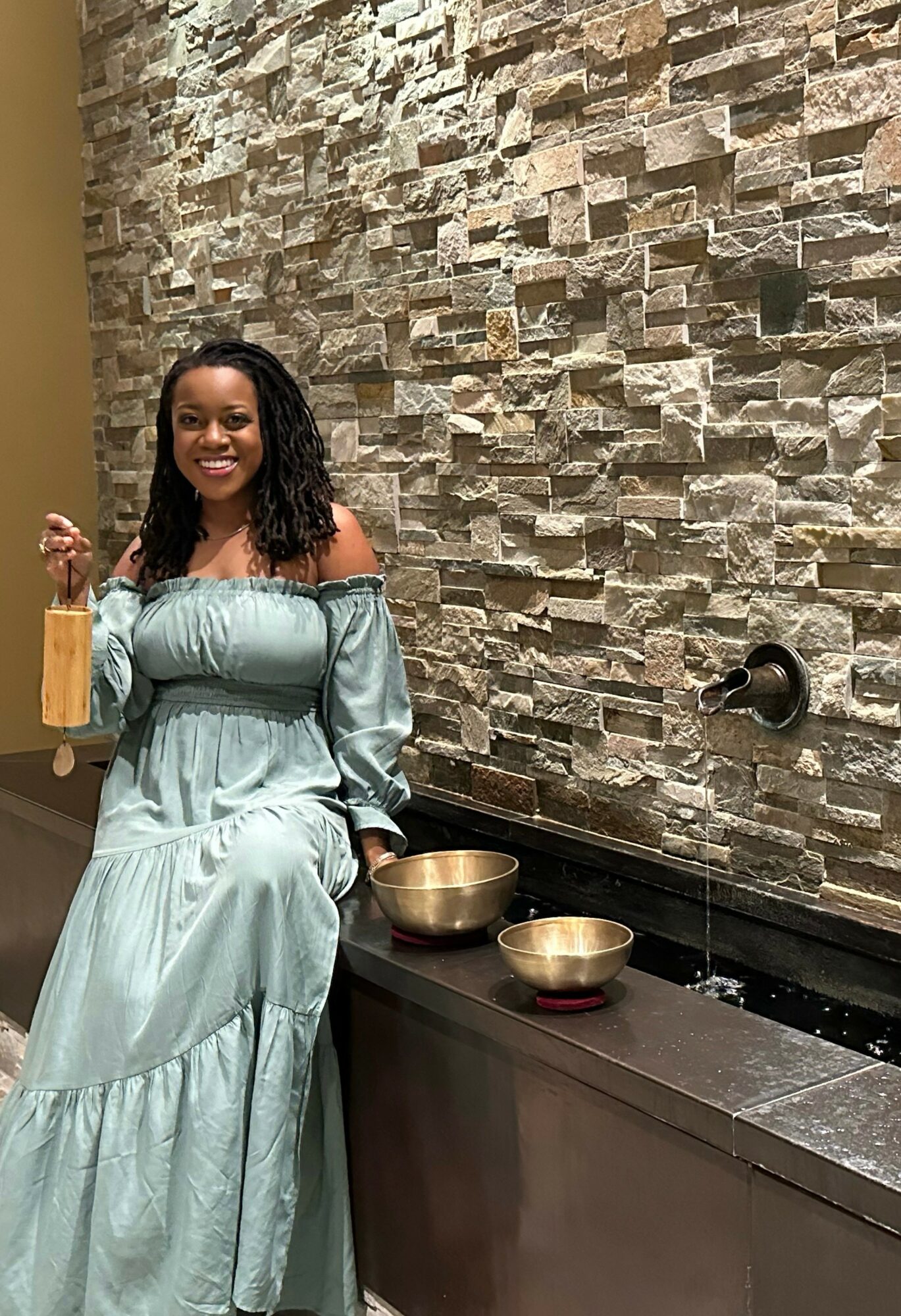 Woman in light blue dress sitting by a stone wall, holding a glass, with bowls on the counter.