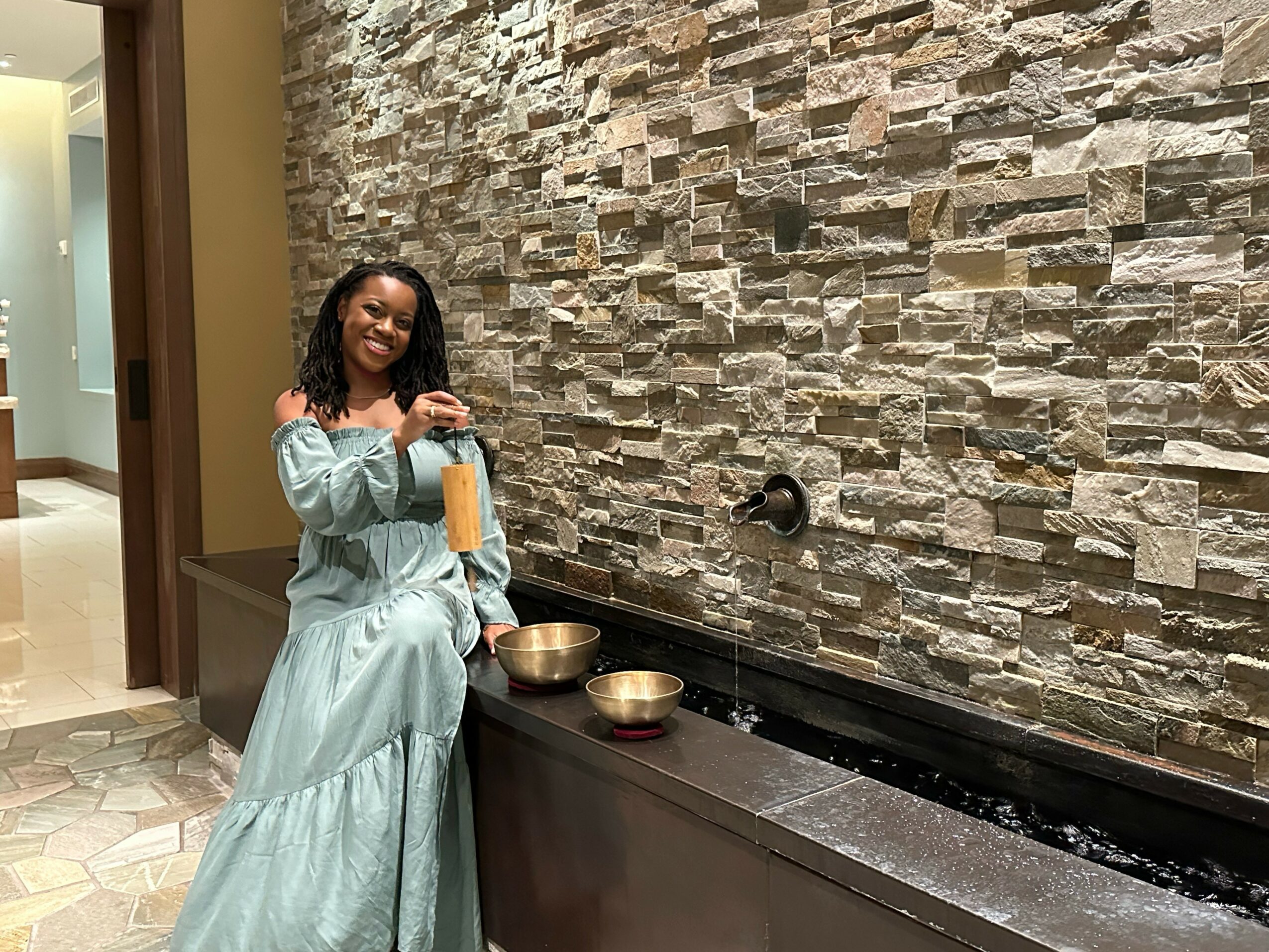 Smiling woman in a light blue dress standing next to a water feature with bowls on a ledge.