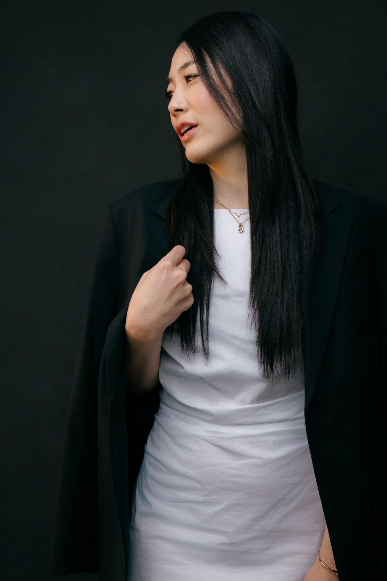 Woman with long black hair wearing a white top and black blazer against dark background.