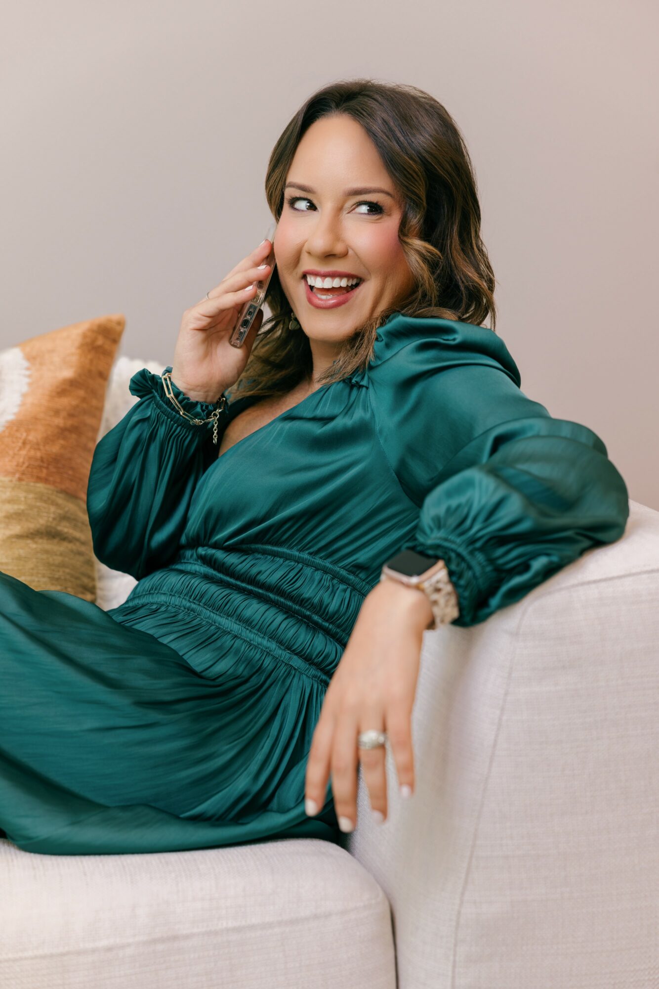 Woman sitting on a sofa, smiling, wearing a teal dress, with a pillow behind her.