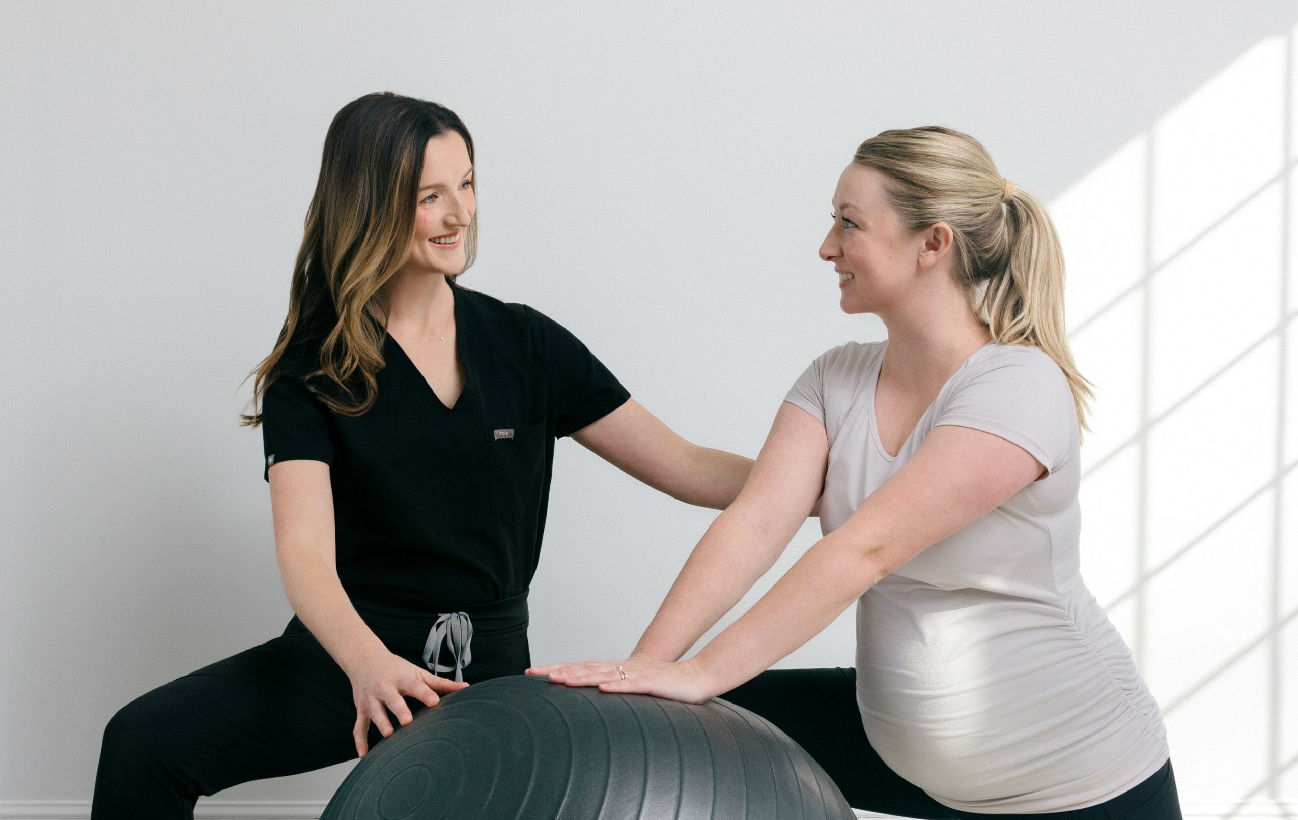 Two women in a clinical setting, one in black scrubs and the other in a white shirt, engaging in a physical therapy session.