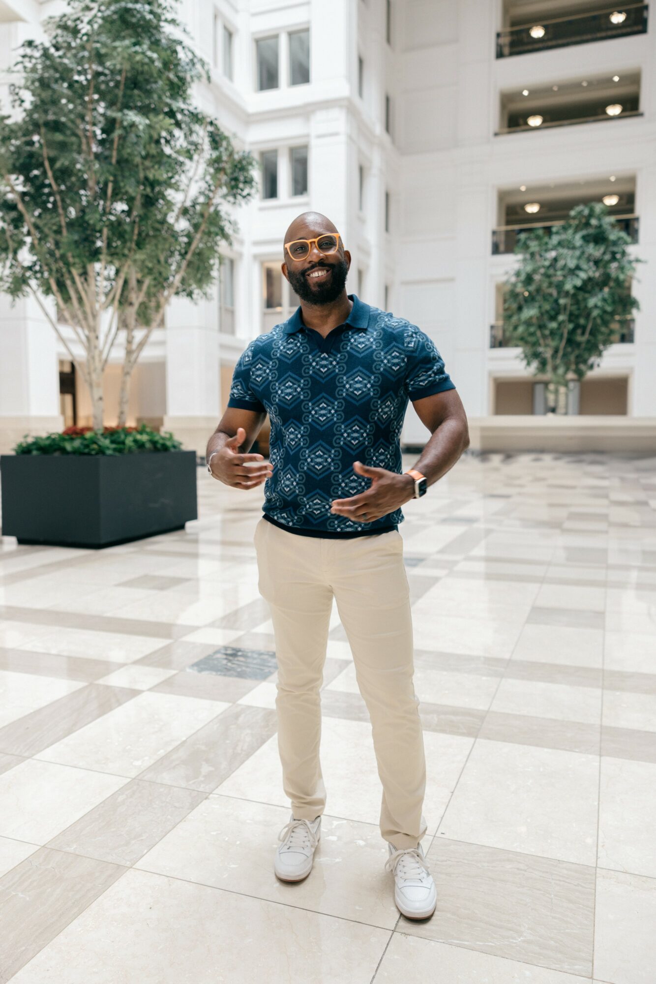Man with glasses and beard smiling, holding a phone and peace sign, standing in a spacious modern building atrium.
