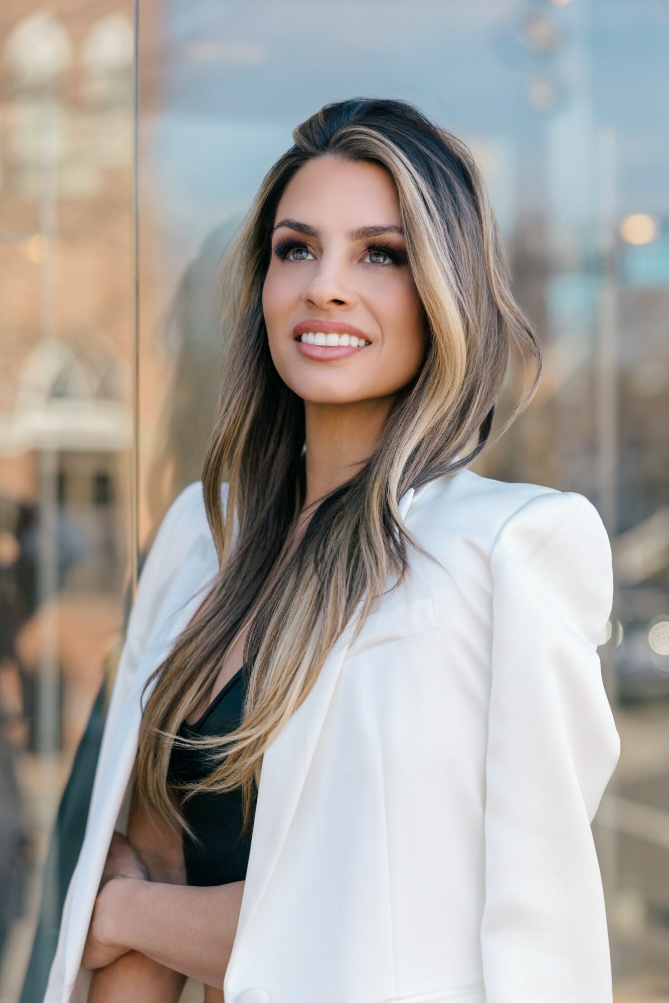 Young woman with long hair and a white blazer smiling outdoors.