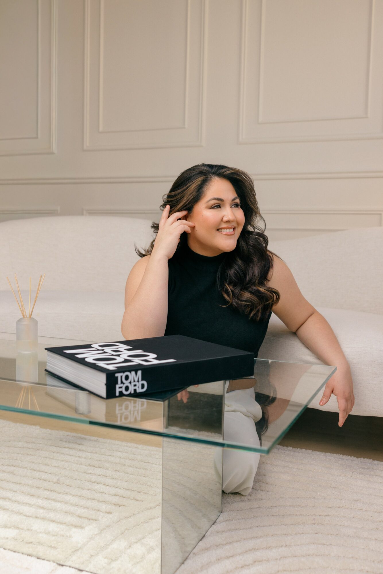 Woman with long dark hair smiling, sitting at a glass table with a black and white book, in a room with beige walls.